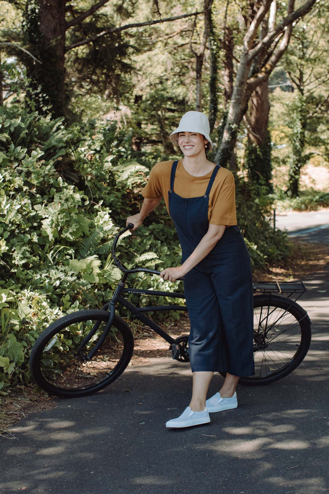 A smiling person wearing the Bridge & Burn Edie Overall in Navy, paired with a mustard shirt and white bucket hat, stands holding a black bicycle on a sunlit path surrounded by green foliage.