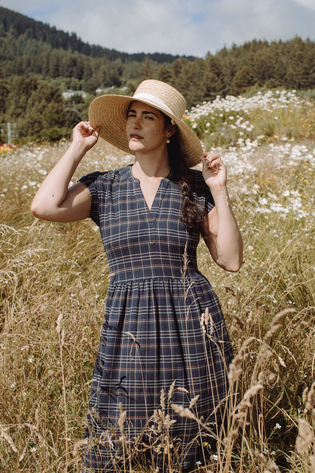 Wearing the Bridge & Burn Ira Dress in Dusk Herringbone Plaid and a wide-brimmed straw hat, a woman stands in tall grass and wildflowers, holding her hat with both hands as trees and hills rise under a partly cloudy sky.