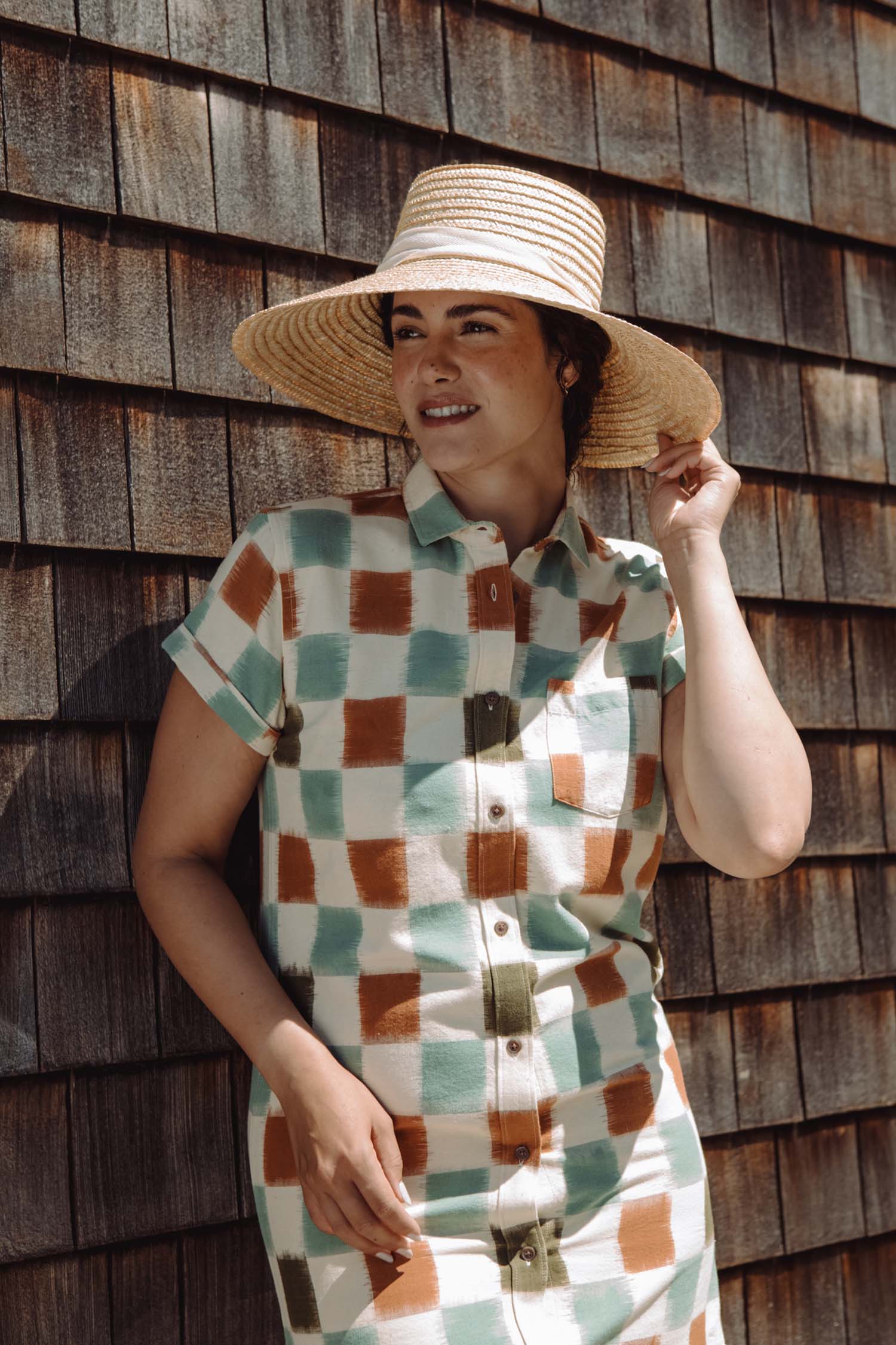 A person wearing the Bridge & Burn Loren Shirt Dress in Mod Checker, a short-sleeve 100% cotton button-front dress with a green, brown, and white check pattern, stands smiling against a wooden shingle wall.