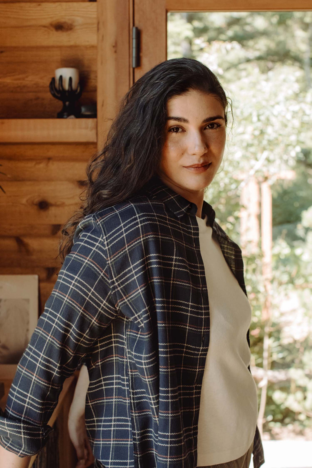 A woman with long dark hair wears the Bridge & Burn Olivia Slim Shirt in Dusk Herringbone Plaid over a white top, standing indoors by a wooden wall and window. She looks at the camera with a slight smile as sunlight and greenery appear outside.