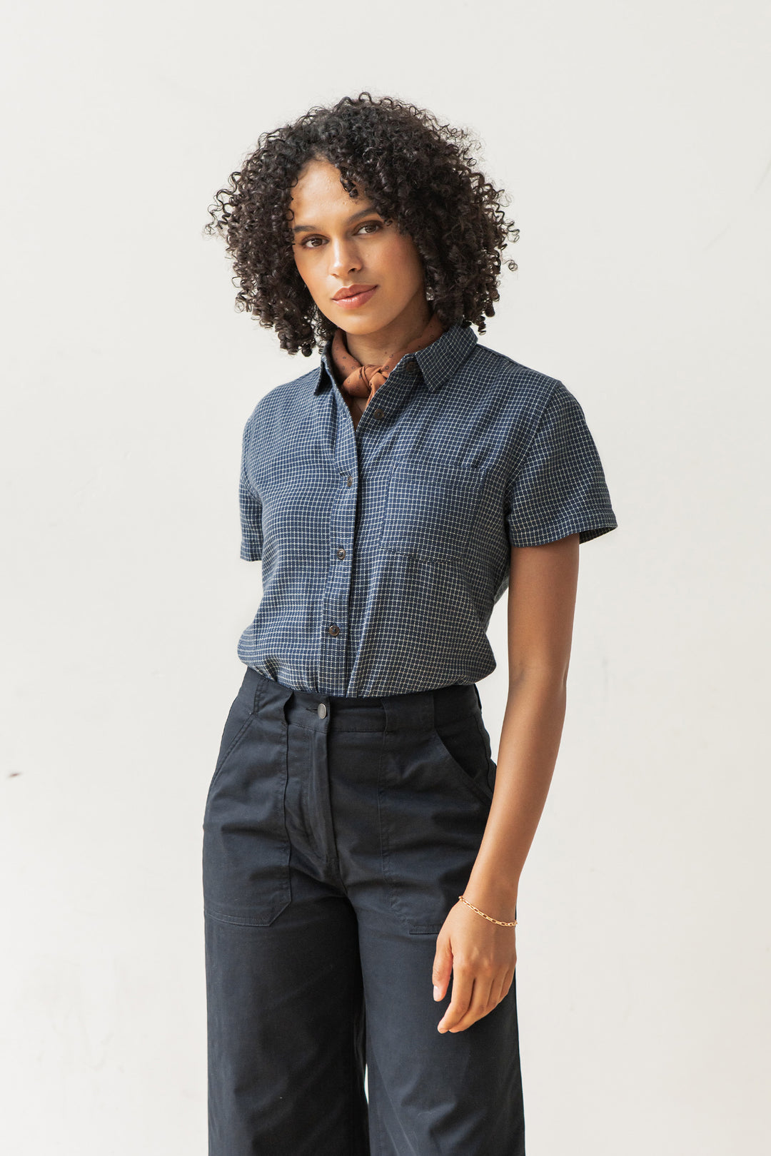 A woman with curly hair wears the Bridge & Burn Lana Shirt in Navy Grid and high-waisted black pants, standing against a plain white background and looking at the camera with a neutral expression.