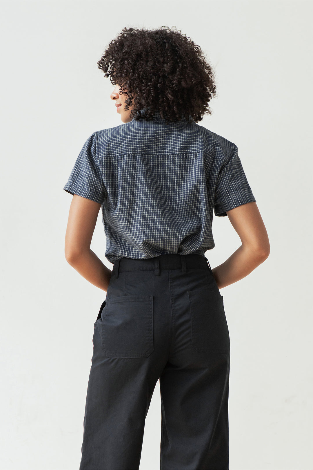 A person with curly hair, seen from the back, wears the Bridge & Burn Lana Shirt in Navy Grid—a 100% cotton, short sleeve button-up—tucked into high-waisted black pants. The background is plain and light-colored.
