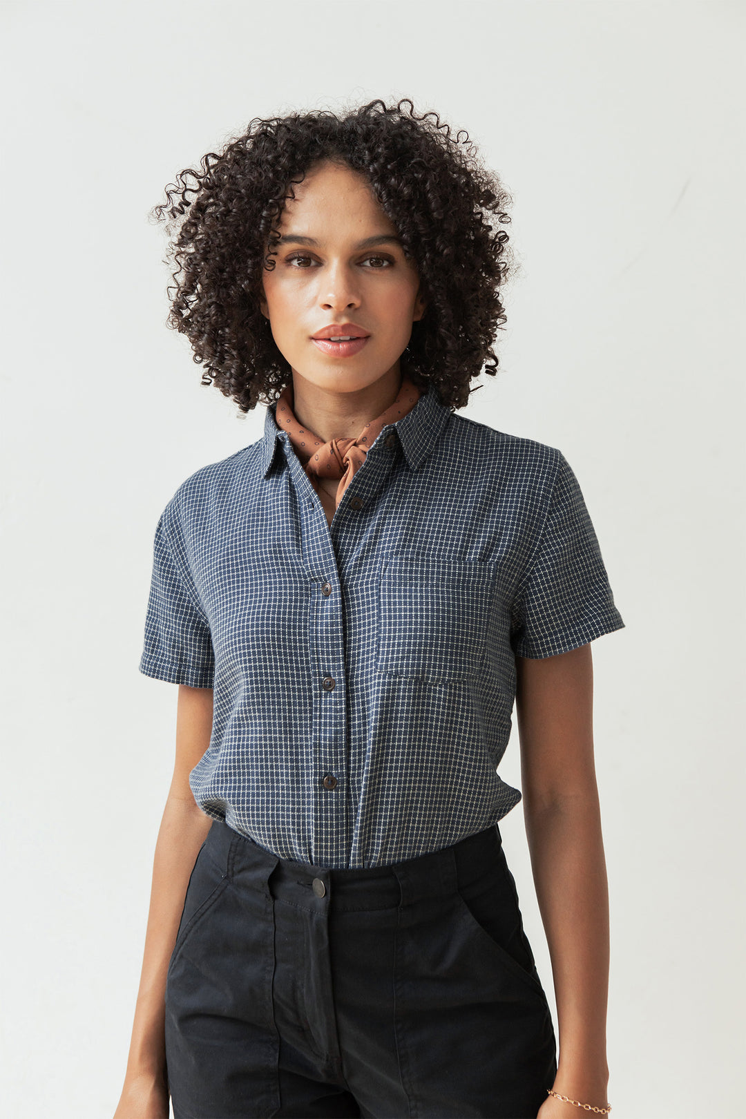 A woman with curly hair wears the Bridge & Burn Lana Shirt in Navy Grid, tucked into dark pants with a light brown neck scarf, standing against a plain light background.