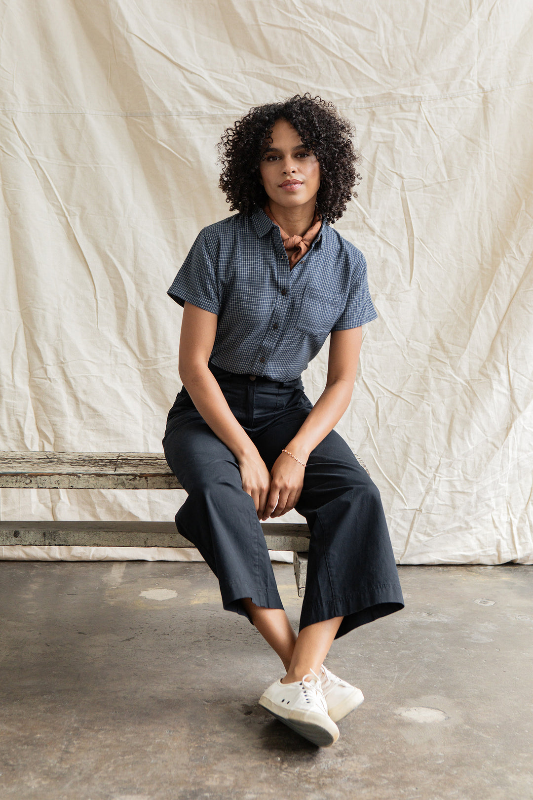 A woman with curly hair sits on a rustic bench, wearing the Bridge & Burn Lana Shirt in Navy Grid, black pants, white sneakers, and a brown scarf, with a beige fabric backdrop behind her.