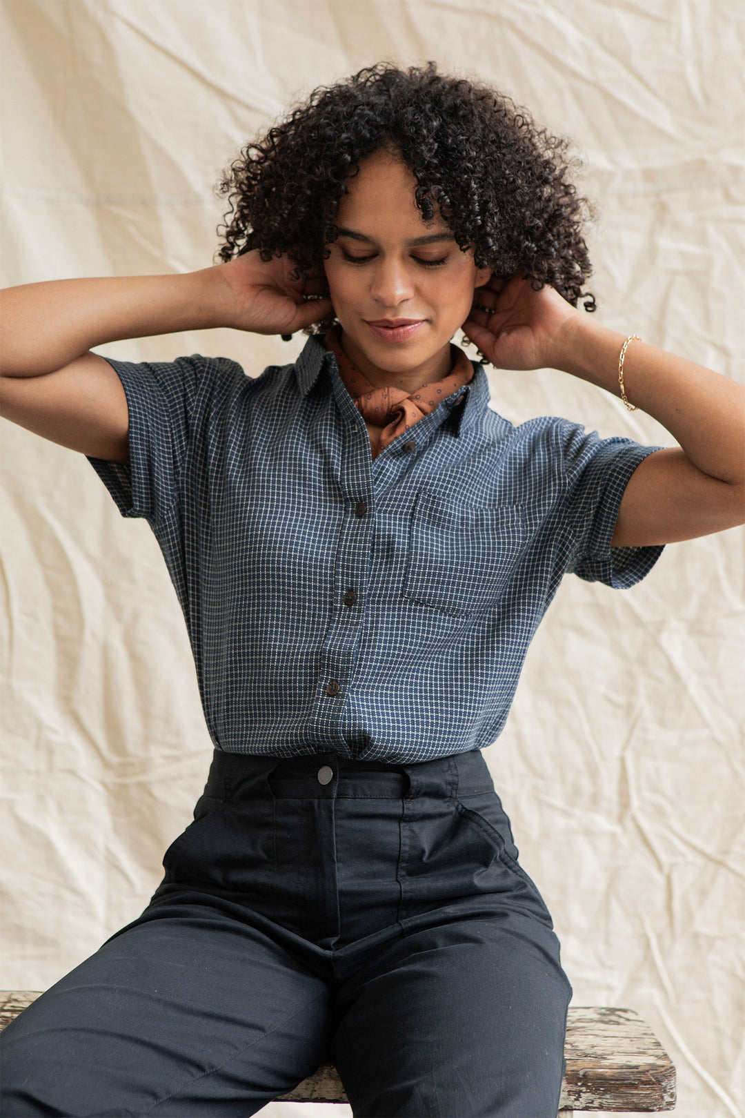 Wearing the Bridge & Burn Lana Shirt in Navy Grid, a person with curly hair and high-waisted black pants sits on a bench adjusting a brown scarf, set against a light fabric backdrop.