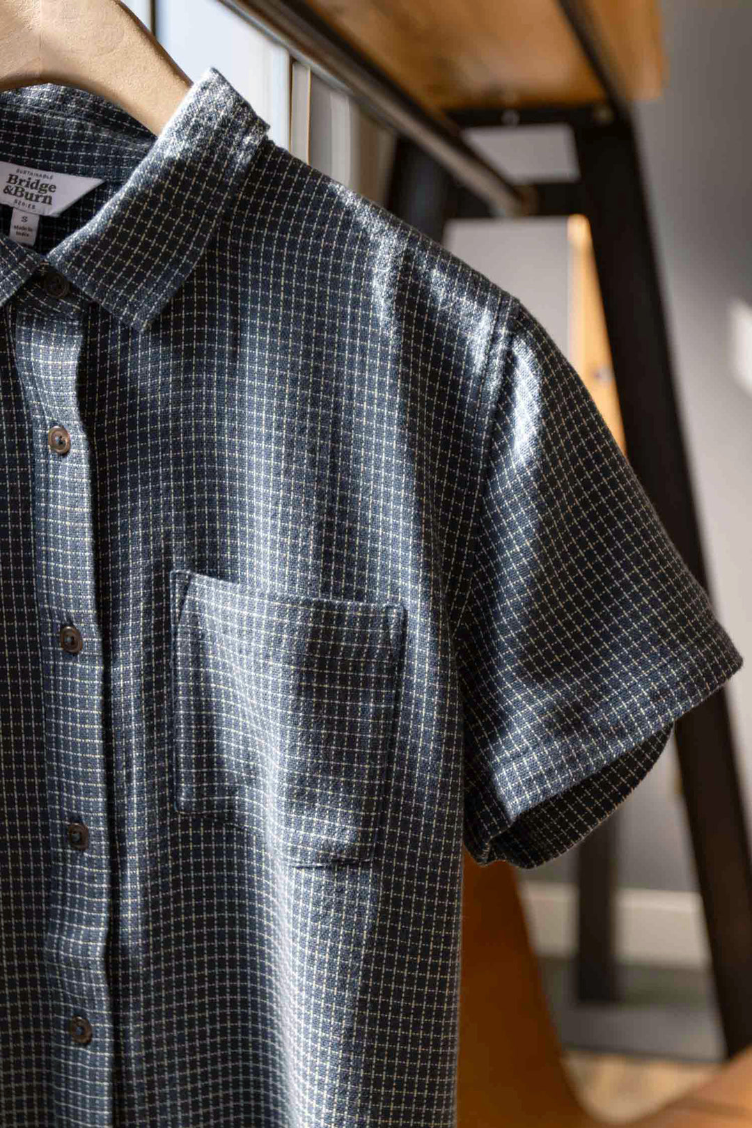 The Bridge & Burn Lana Shirt in Navy Grid, a relaxed fit short sleeve button-up made from 100% cotton with a chest pocket and brown buttons, hangs on a wooden hanger in a sunlit room.