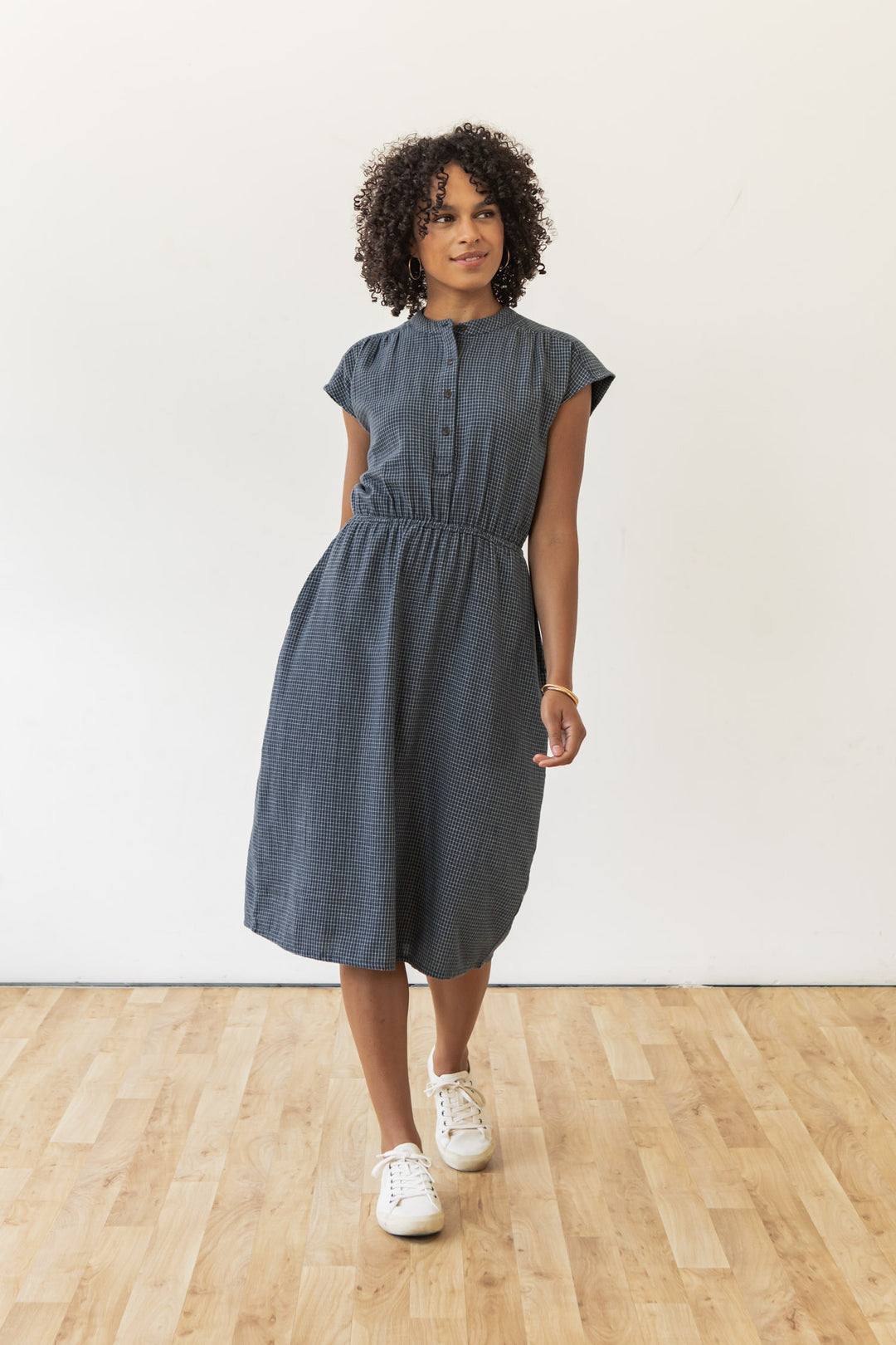 A woman with curly hair wears the Bridge & Burn Lorane Dress in Navy Grid, a casual short-sleeve A-line style with buttons and an elastic waist. The knee-length dress pairs perfectly with white sneakers as she stands by a plain white wall.