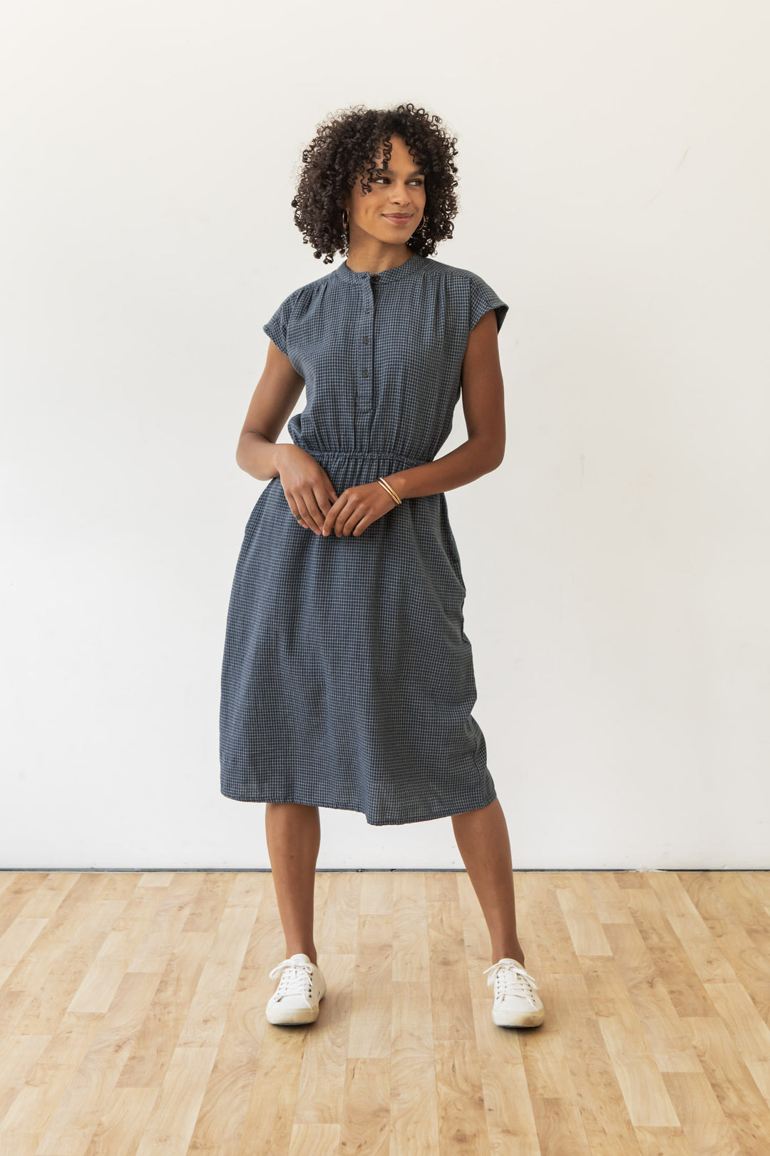 A woman with curly hair wears the Lorane Dress in Navy Grid by Bridge & Burn and white sneakers, standing on a wooden floor against a white wall, smiling slightly with her hands at her waist.