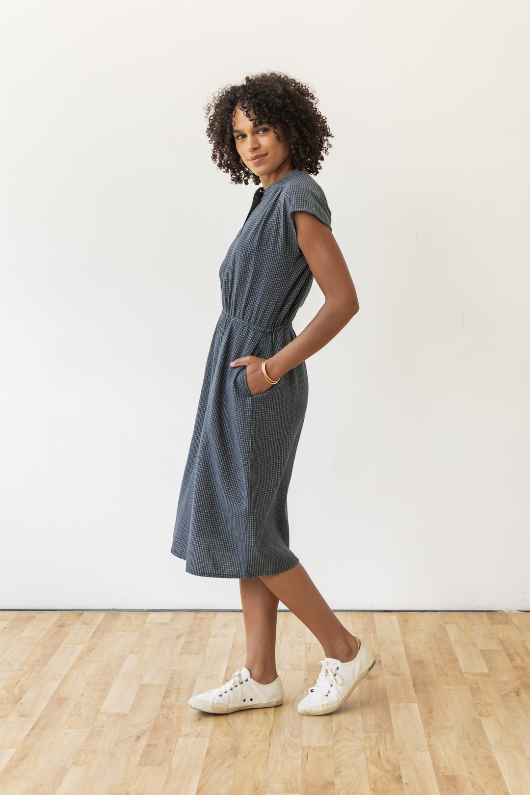 A woman with curly hair wears the Lorane Dress in Navy Grid by Bridge & Burn—a casual, short-sleeve, textured cotton A-line dress with pockets—paired with white sneakers. She stands on a wooden floor against a plain white wall, looking relaxed.