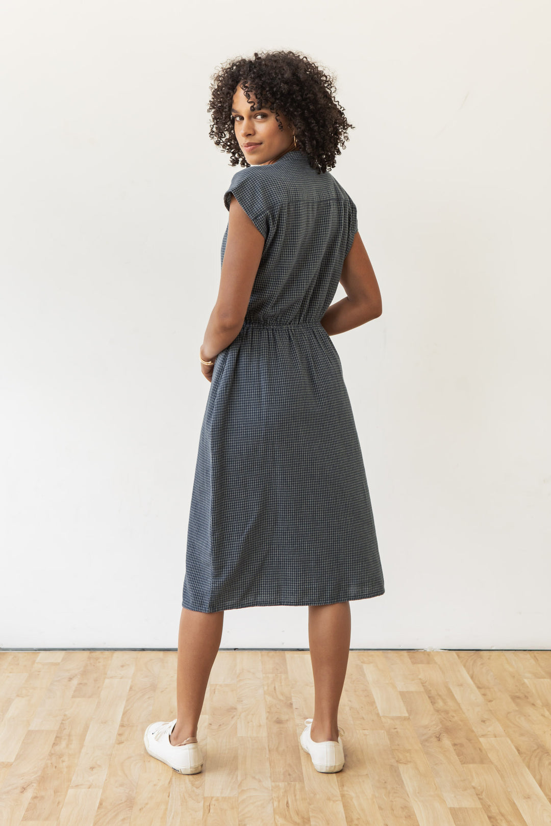 A woman with curly hair stands on a wooden floor, glancing over her shoulder. She wears the Bridge & Burn Lorane Dress in Navy Grid with white sneakers, set against a plain white wall.