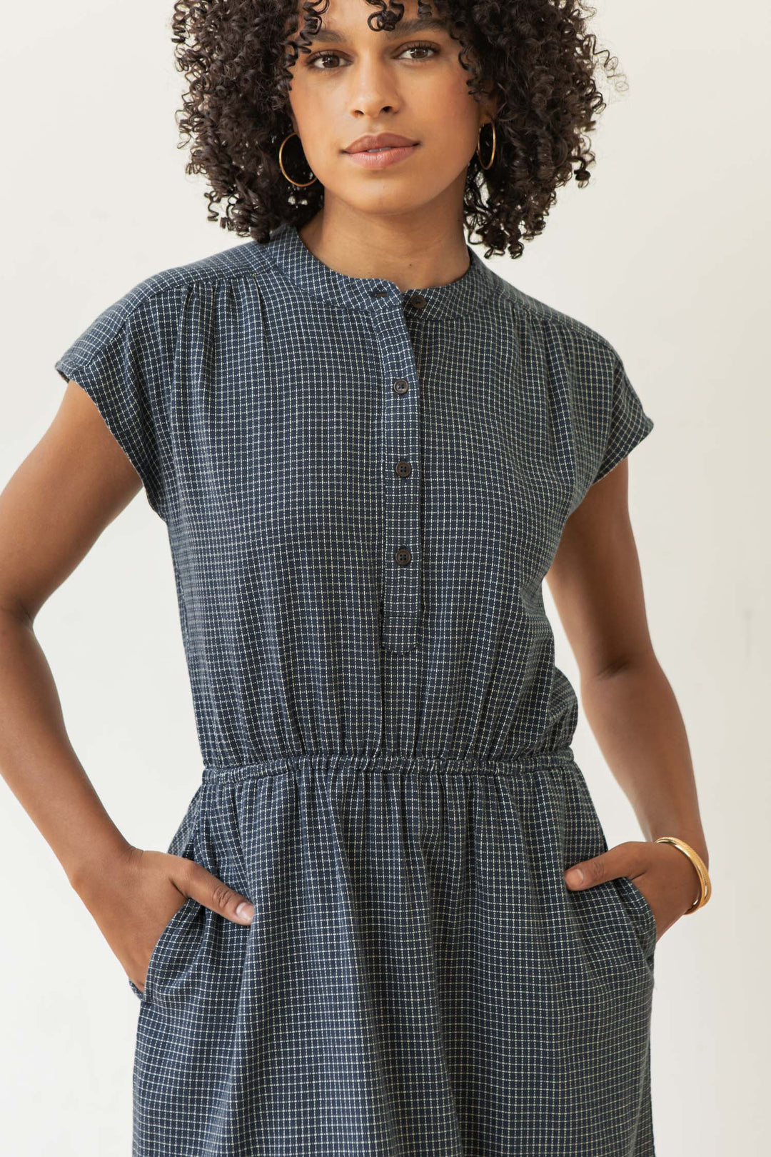 A woman with curly hair wears the Bridge & Burn Lorane Dress in Navy Grid—an A-line cotton dress with buttons, an elastic waist, and pockets. She stands against a plain background, accessorized with hoop earrings and a bracelet.