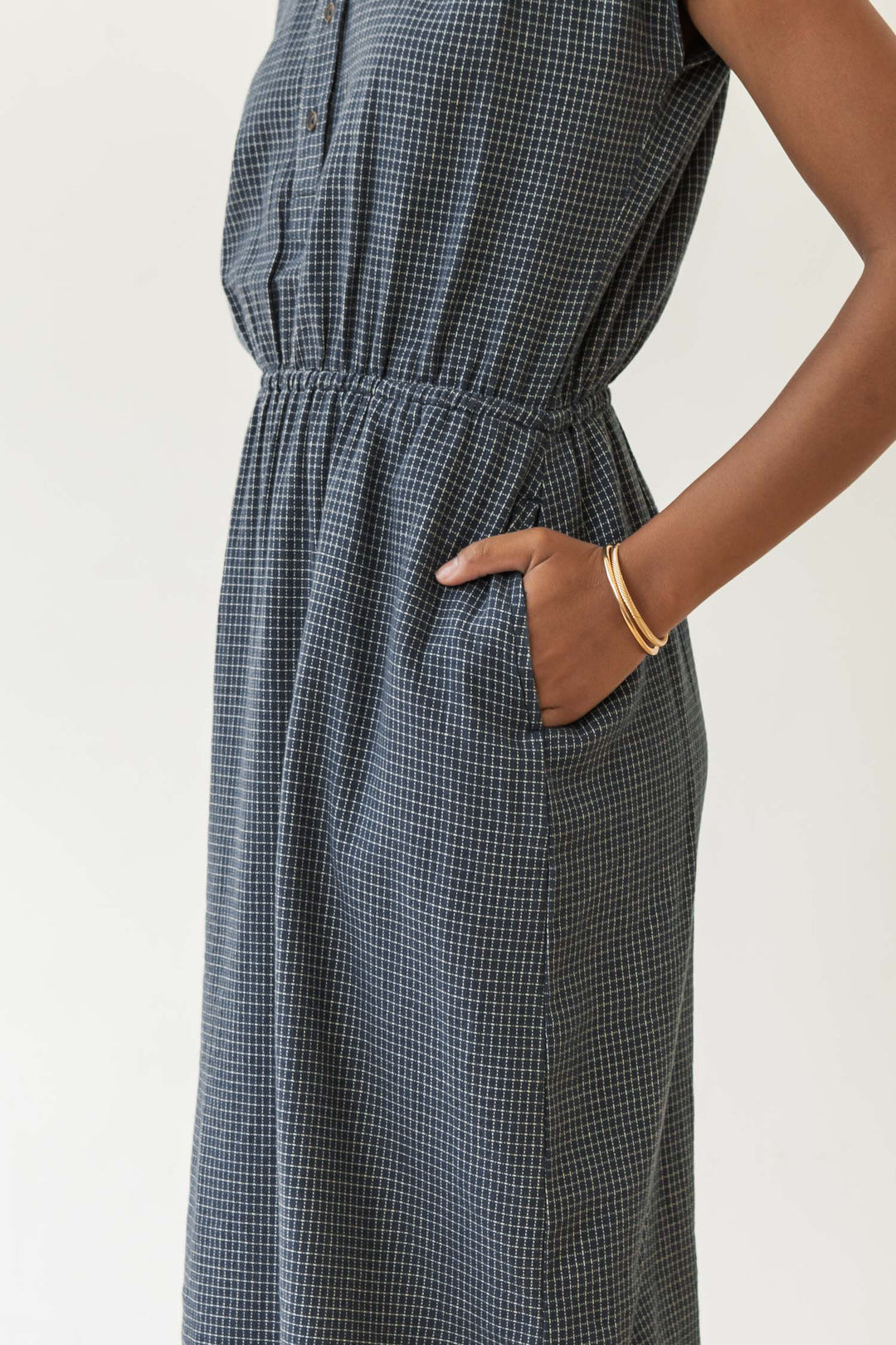 A person wears the Bridge & Burn Lorane Dress in Navy Grid, a sleeveless cotton dress with an elastic waistband and side pocket, styled with gold bangles against a plain light background.