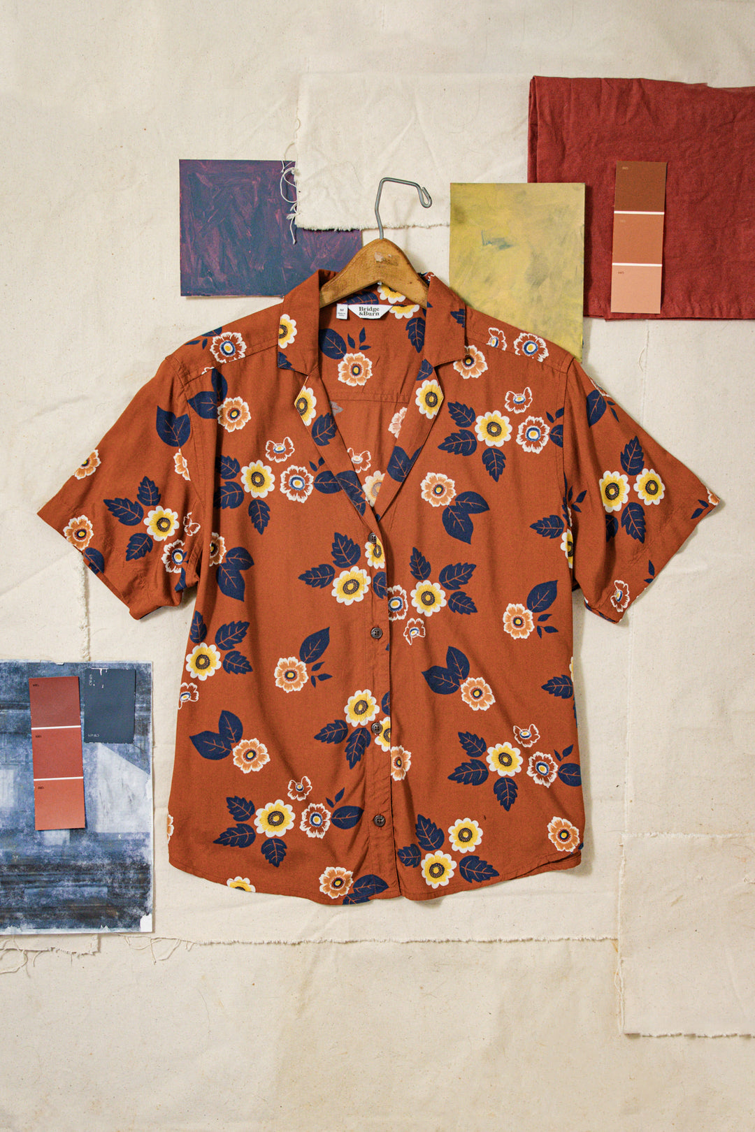 The Bridge & Burn Mila Cropped Shirt in Rose Floral, a relaxed fit short-sleeve button-up with yellow, white, and blue floral leaf patterns on a rust background, hangs on a wooden hanger amid textured fabrics and paint swatches.
