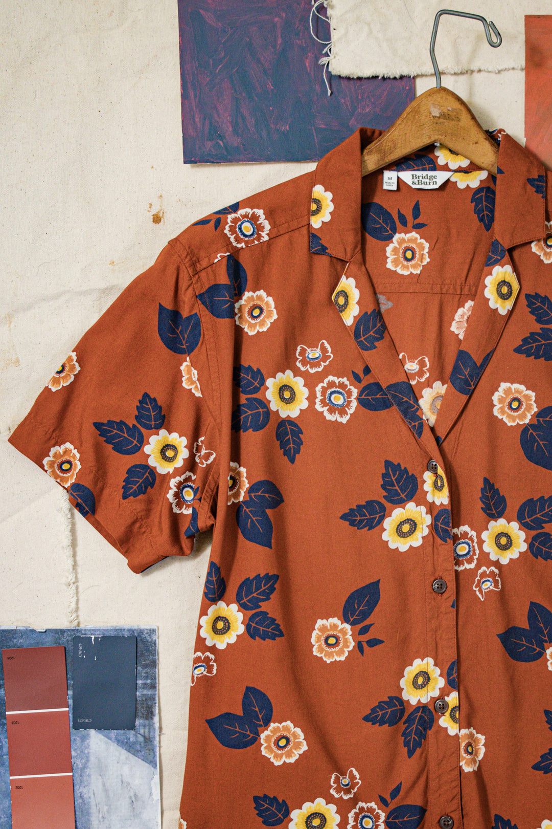 The Bridge & Burn Mila Cropped Shirt in Rose Floral hangs on a wooden hanger against a textured off-white wall with color swatches and paper in the background.