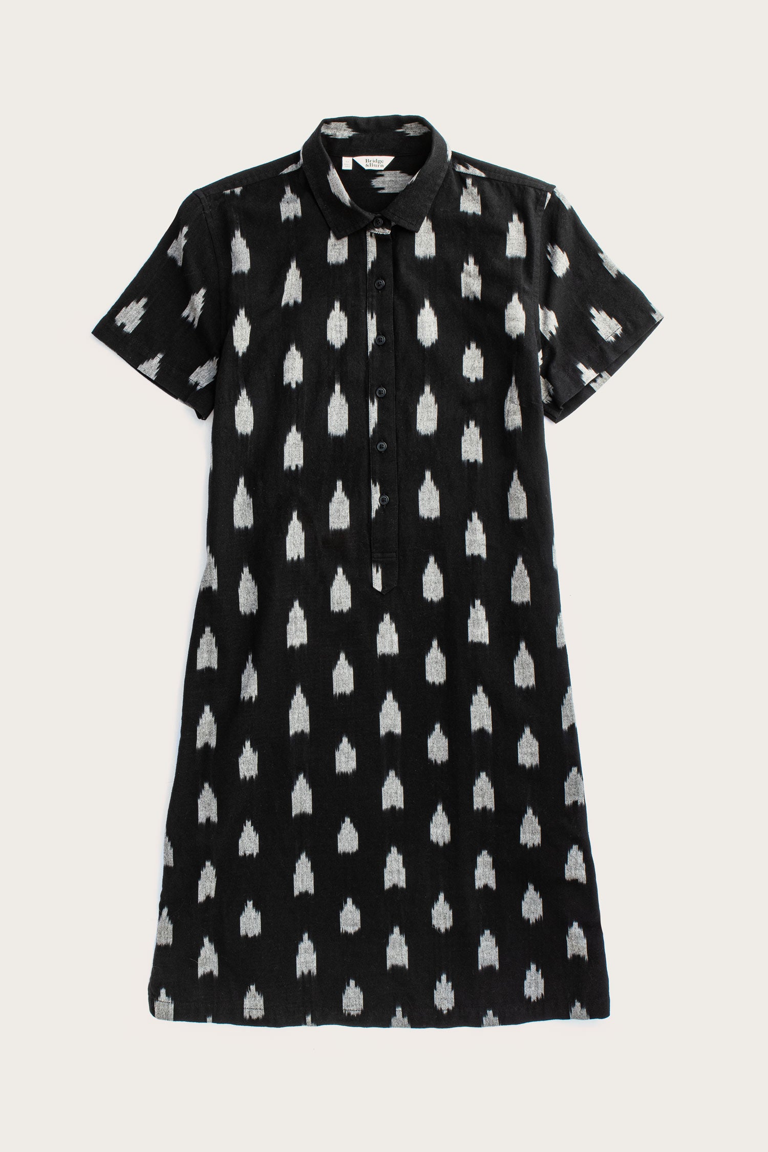 The Delia Shirt Dress in Eclipse Ikat by Bridge & Burn is a knee-length, black organic cotton dress with short sleeves, a collar, front buttons, and an allover white ikat pattern.