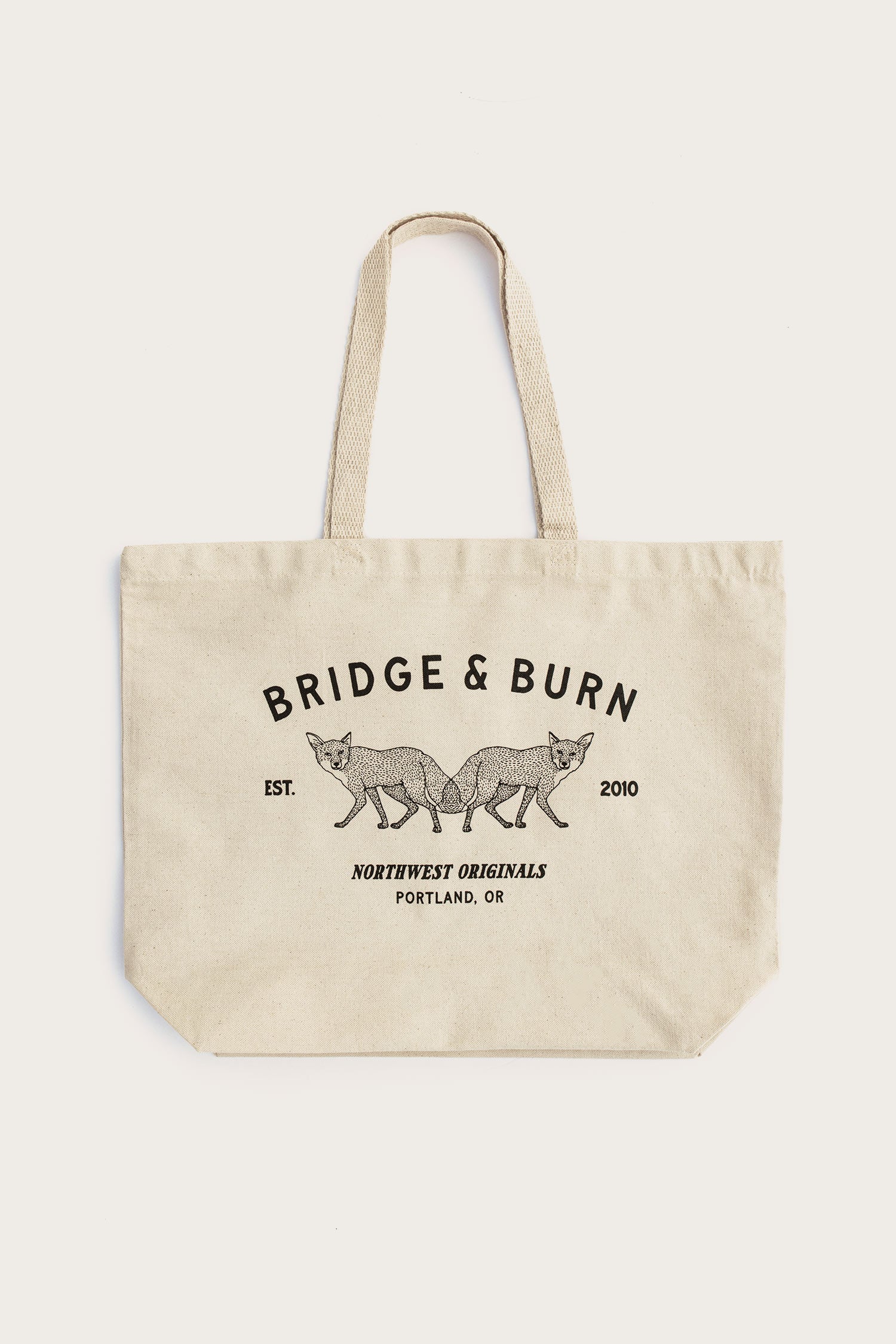 The Fox Tote / Natural by Bridge & Burn is a beige cotton canvas tote with two handles, featuring BRIDGE & BURN and twin foxes artwork. It says "EST. 2010" and "NORTHWEST ORIGINALS PORTLAND, OR." Proudly made in the USA.