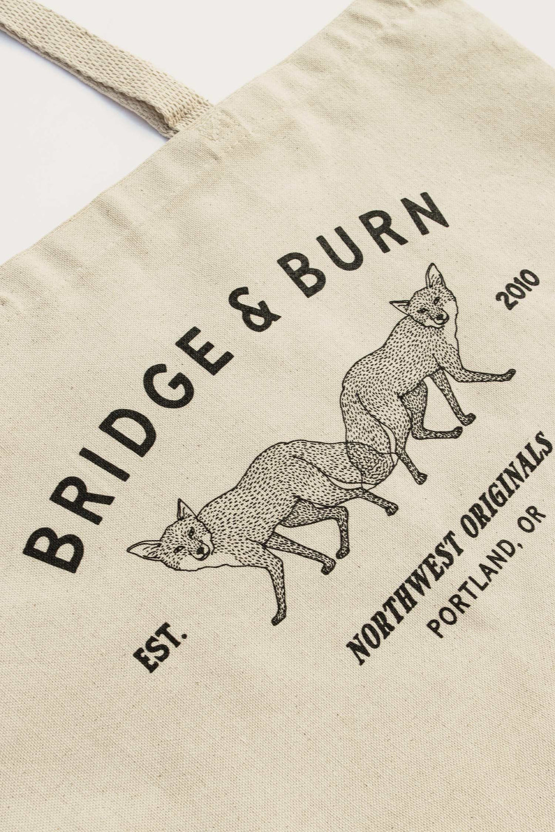 The Bridge & Burn Fox Tote / Natural is a beige cotton canvas bag featuring two illustrated foxes and black text: "BRIDGE & BURN, Est. 2010, Northwest Originals, Portland, OR.