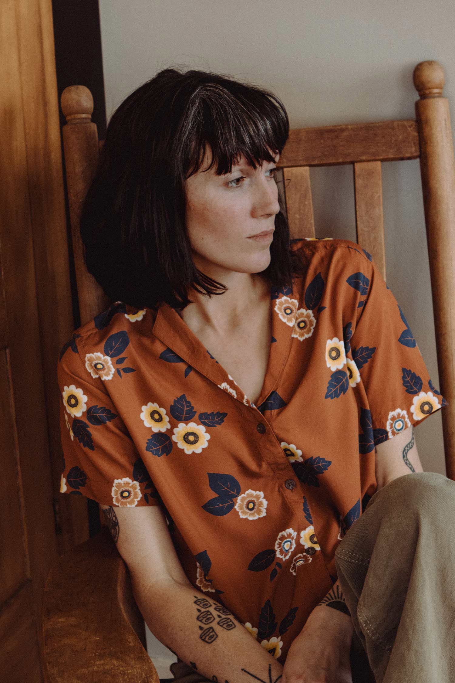 A woman with shoulder-length dark hair sits on a wooden chair, wearing Bridge & Burn's Mila Cropped Shirt in Rose Floral. She gazes thoughtfully to the side, embodying the relaxed style of this floral top.