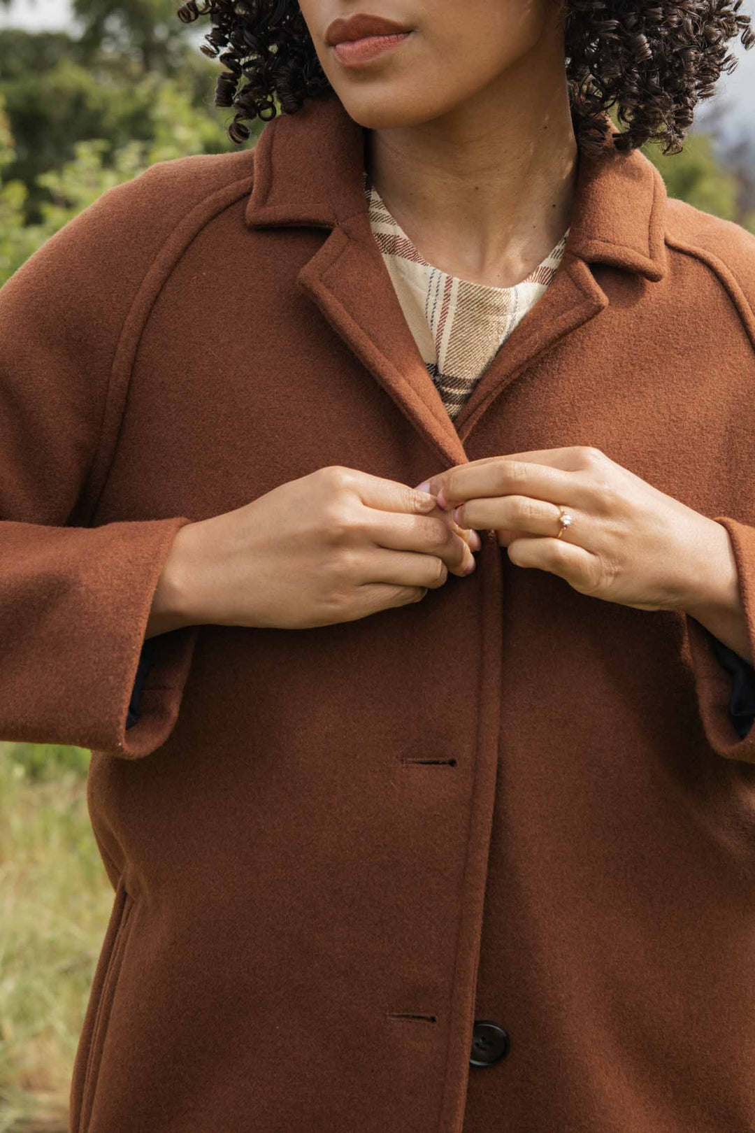 A person with curly hair fastens the buttons on a Bridge & Burn Alberta Car Coat in Fired Brick. Outdoors amid greenery, only the lower part of their face is visible.