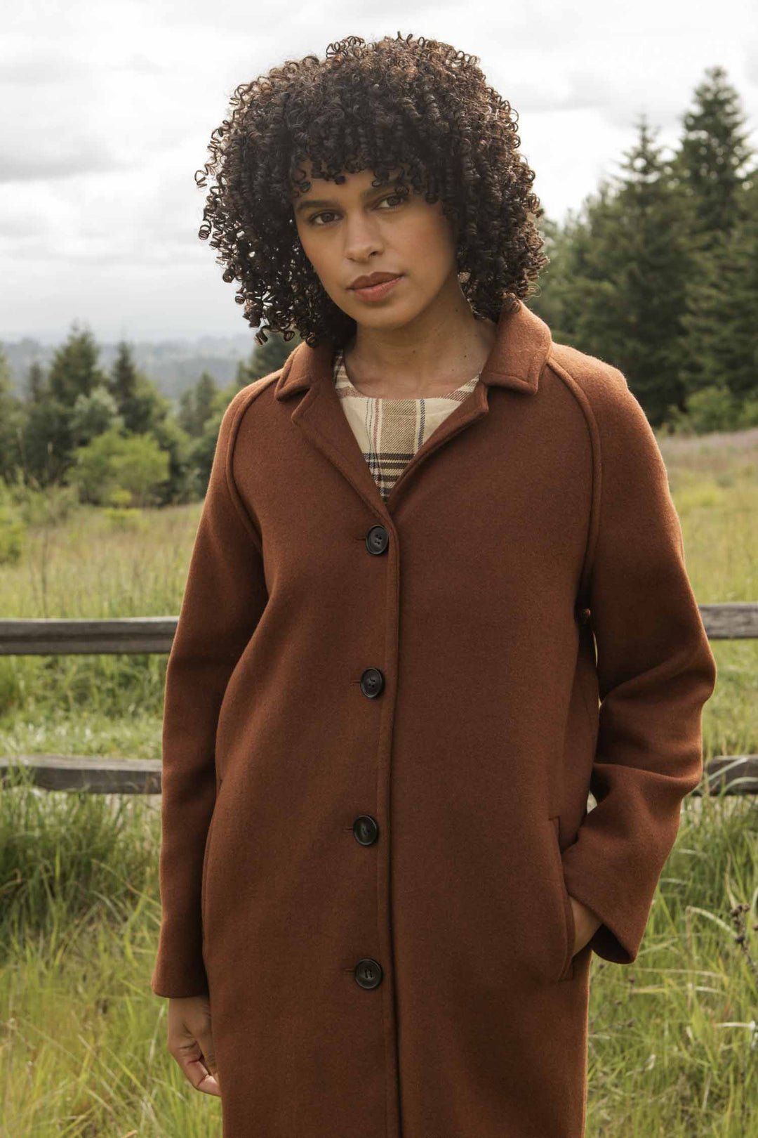 A person with curly hair wears the Bridge & Burn Alberta Car Coat in Fired Brick over a plaid shirt, standing outdoors by a wooden fence with trees and greenery in the background.