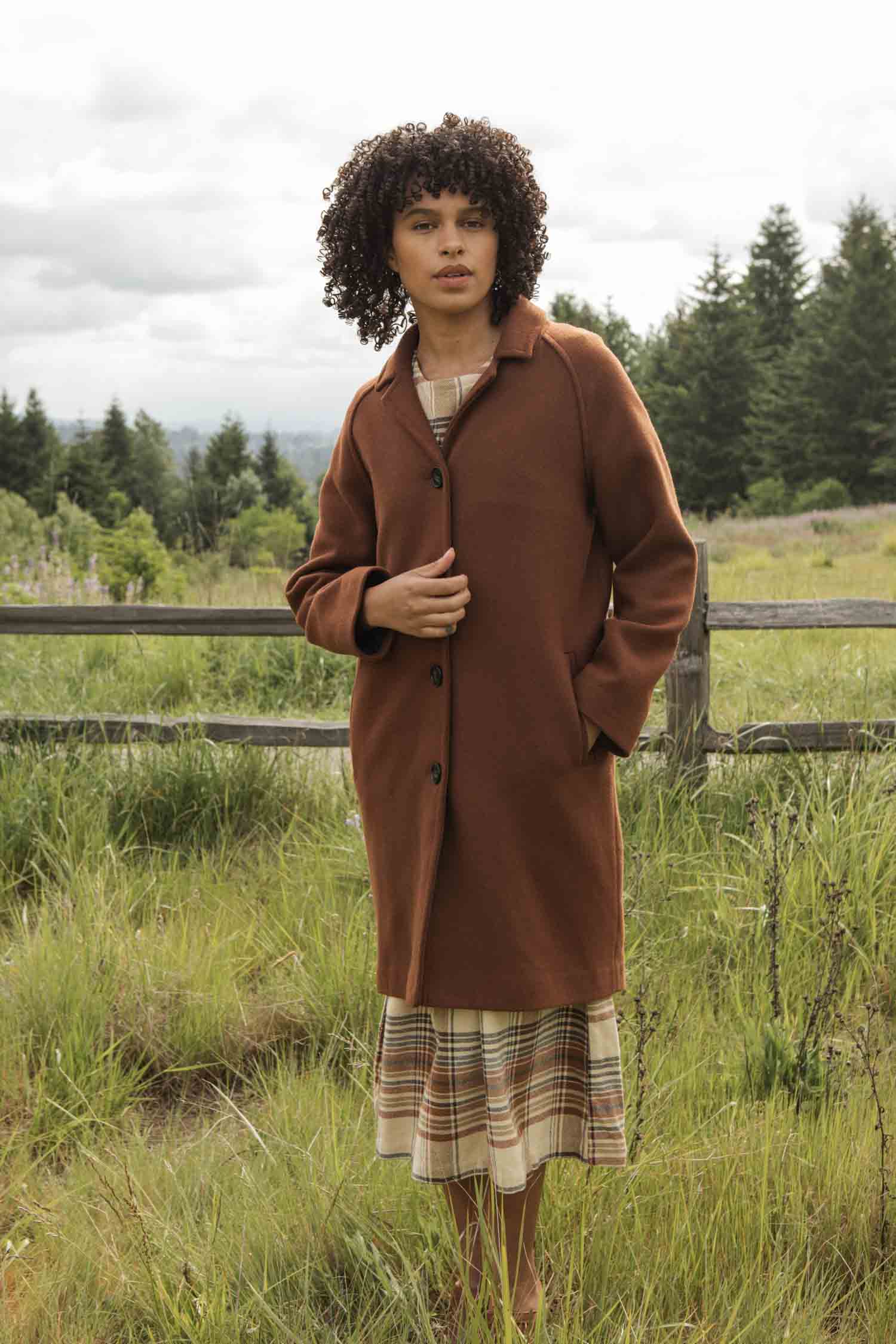 A woman with curly hair stands in a grassy field, wearing the Bridge & Burn Alberta Car Coat in Fired Brick over a plaid dress. Trees and a wooden fence are visible behind her under a cloudy sky.