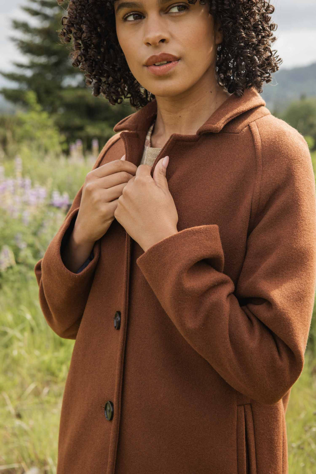 A person with curly hair stands in a field, wearing the Bridge & Burn Alberta Car Coat in Fired Brick and holding the collar with both hands. Greenery and flowers blur in the background.