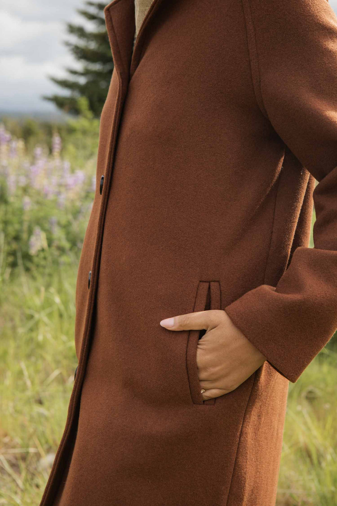 A person wearing the Bridge & Burn Alberta Car Coat in Fired Brick stands outdoors with one hand in a pocket, set against green grass, wildflowers, and a tree for a natural vibe.