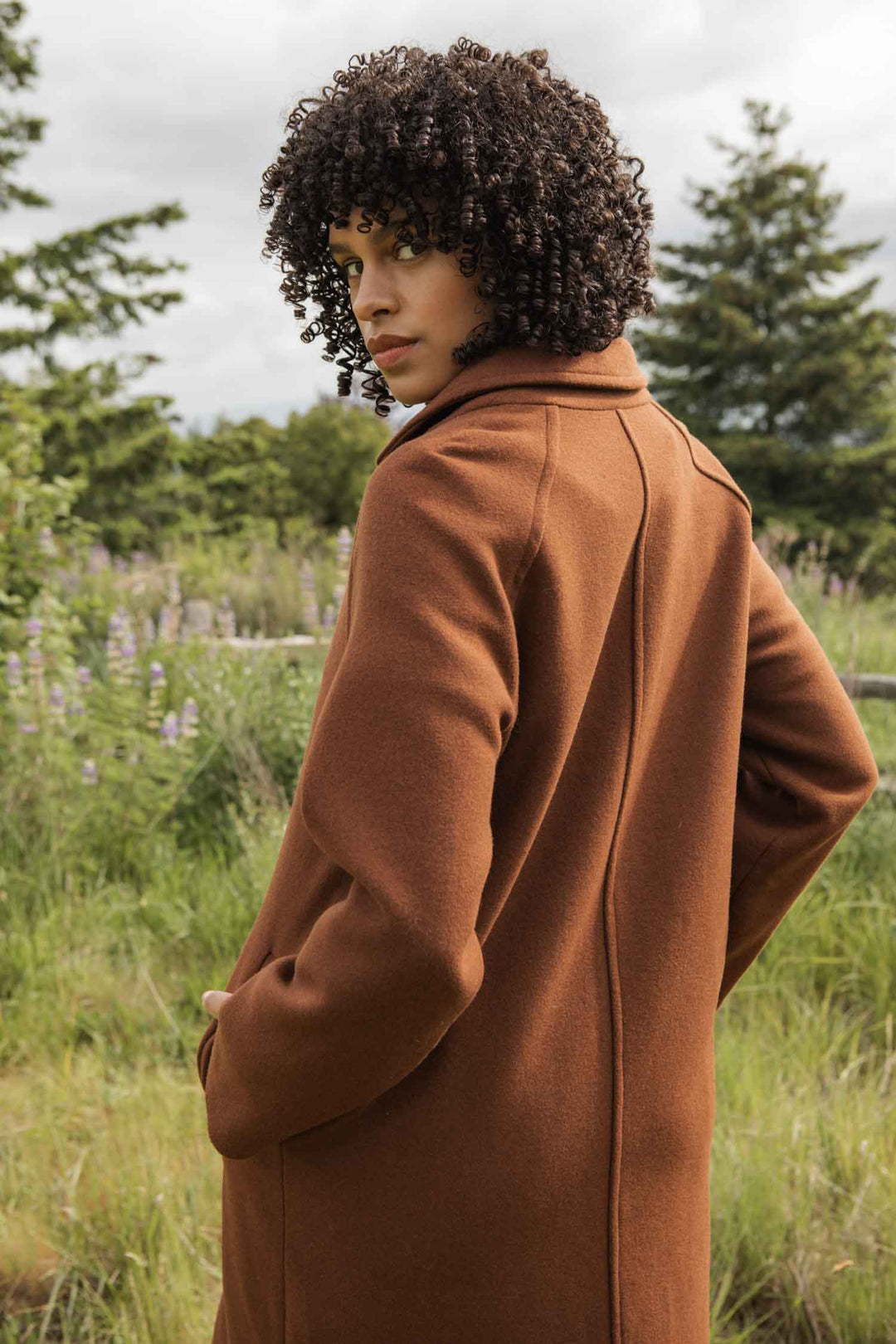 Wearing the Bridge & Burn Alberta Car Coat in Fired Brick, a person with curly hair stands outdoors among grass, trees, and wildflowers, looking over their shoulder at the camera.