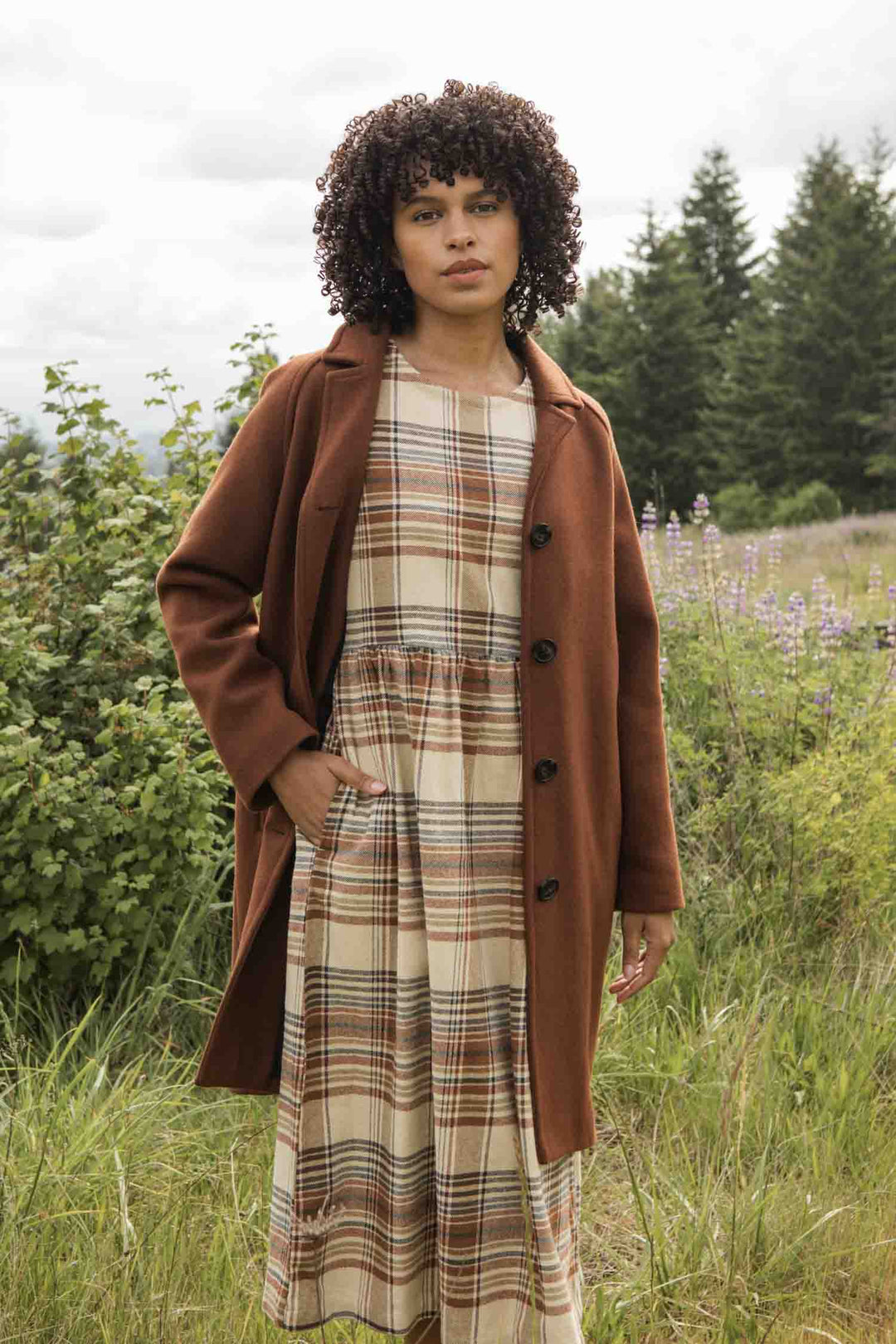 A woman with curly hair stands in a grassy field under a cloudy sky, wearing the Bridge & Burn Alberta Car Coat in Fired Brick over a beige and brown plaid dress. Green trees and plants are visible in the background.