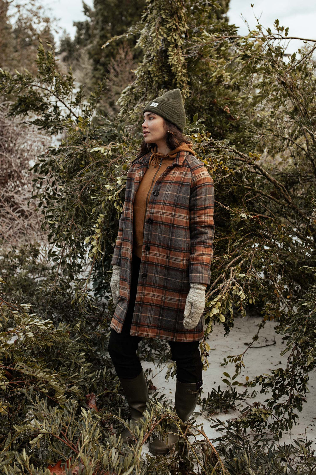 Wearing the Bridge & Burn Alberta Car Coat in Dorset Brushed Plaid, along with a knit hat, gloves, and boots, a person stands among snow-dusted branches and greenery in a wintry outdoor scene.