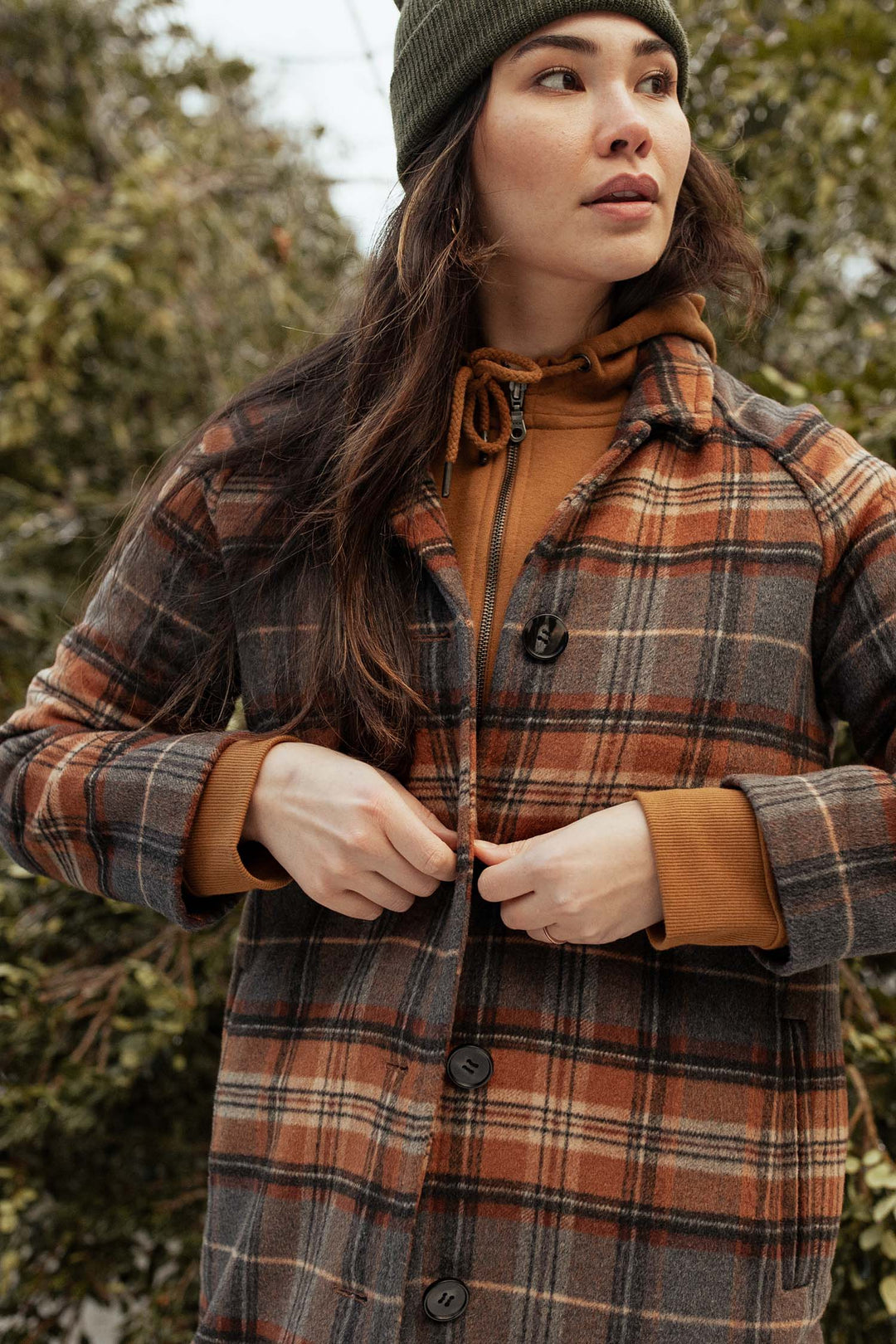 A woman wearing a green knit beanie, brown hoodie, and the Bridge & Burn Alberta Car Coat in Dorset Brushed Plaid stands outdoors by trees, looking to the side as she buttons her heavyweight wool coat.