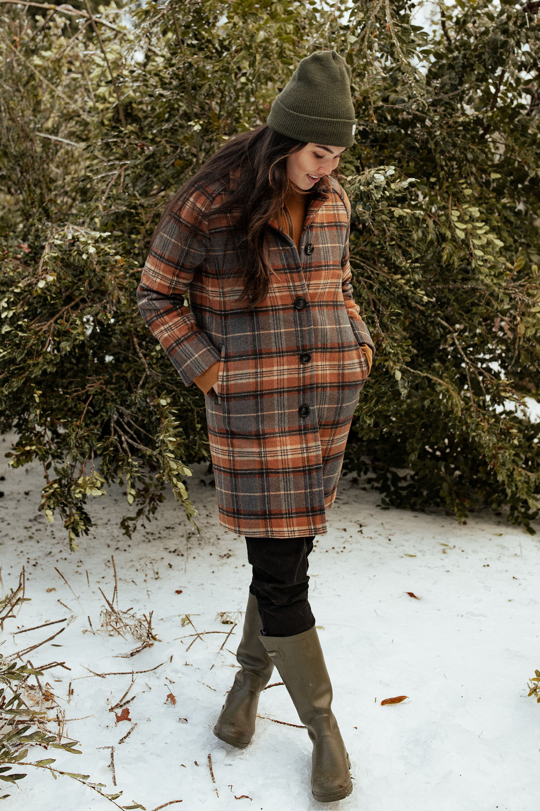 A person wearing the Bridge & Burn Alberta Car Coat in Dorset Brushed Plaid stands on snow with hands in pockets, complemented by a green beanie and boots. Evergreen branches complete the wintry scene.