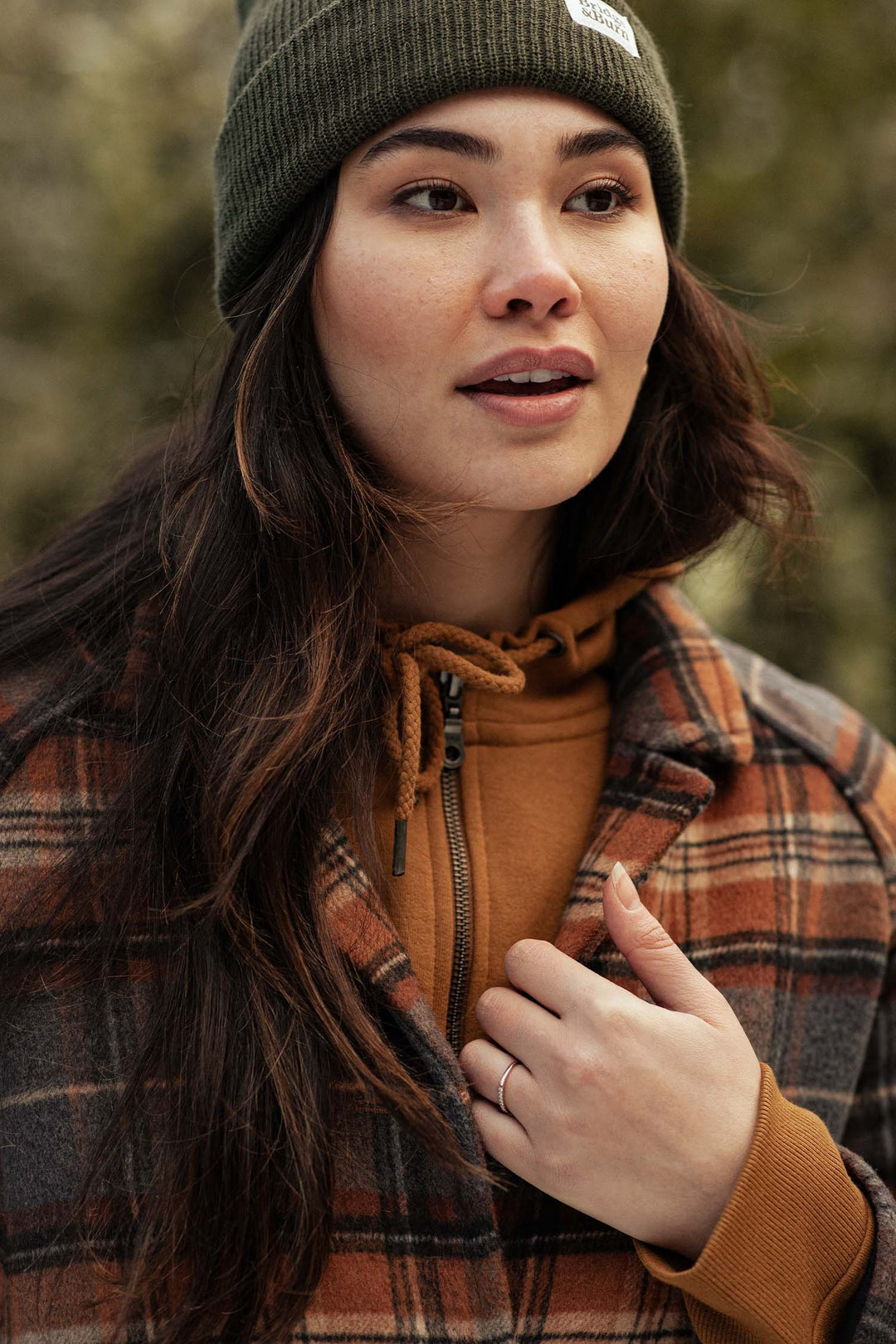A woman with long dark hair wears a green beanie, brown hoodie, and the Bridge & Burn Alberta Car Coat in Dorset Brushed Plaid. She holds the coat collar with one hand, looking slightly to the side against a blurred outdoor background.