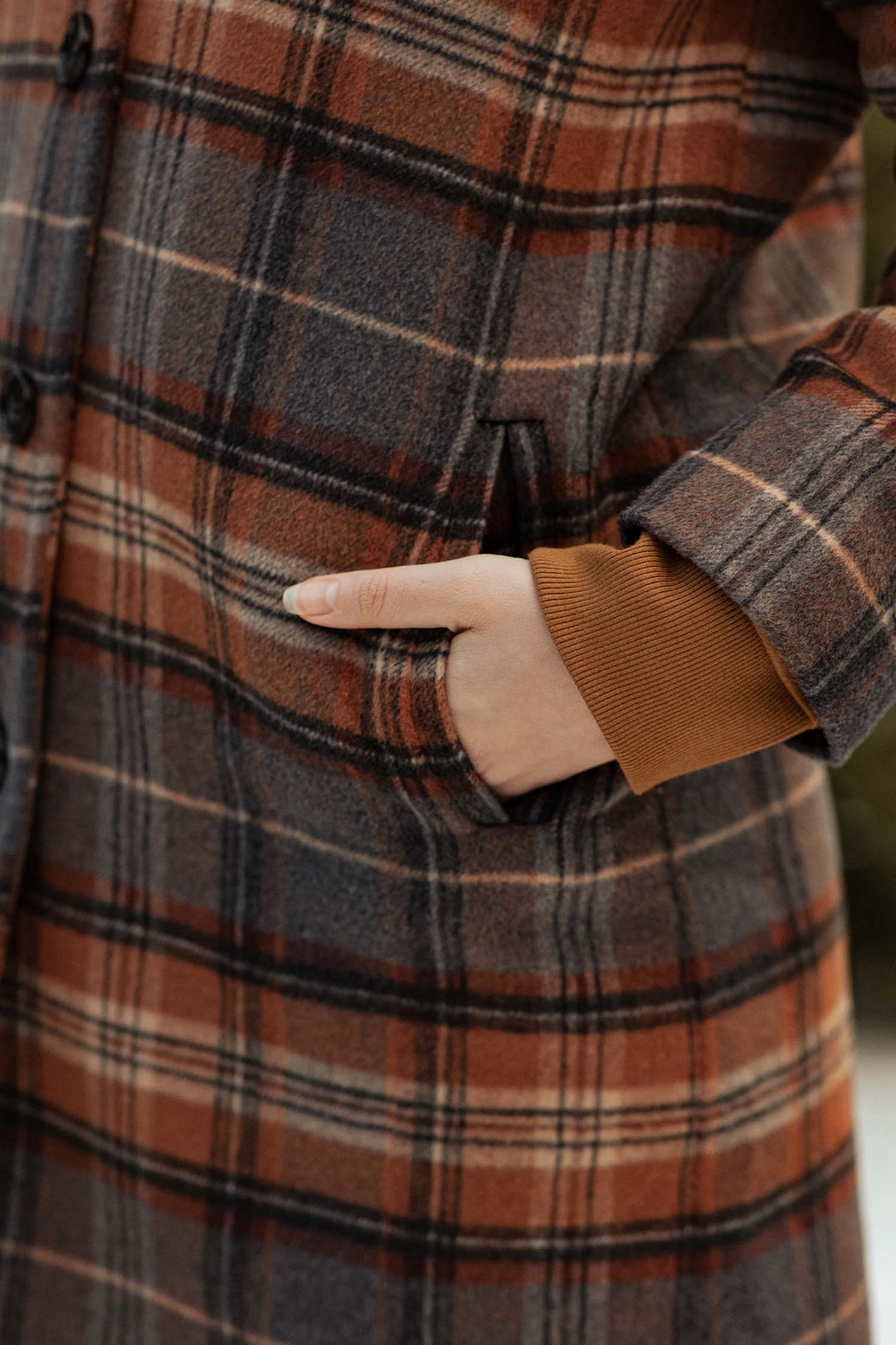 A close-up shows a hand in the pocket of Bridge & Burn’s Alberta Car Coat in Dorset Brushed Plaid—heavyweight wool with brown, orange, and gray tones—worn over a brown long-sleeve top.