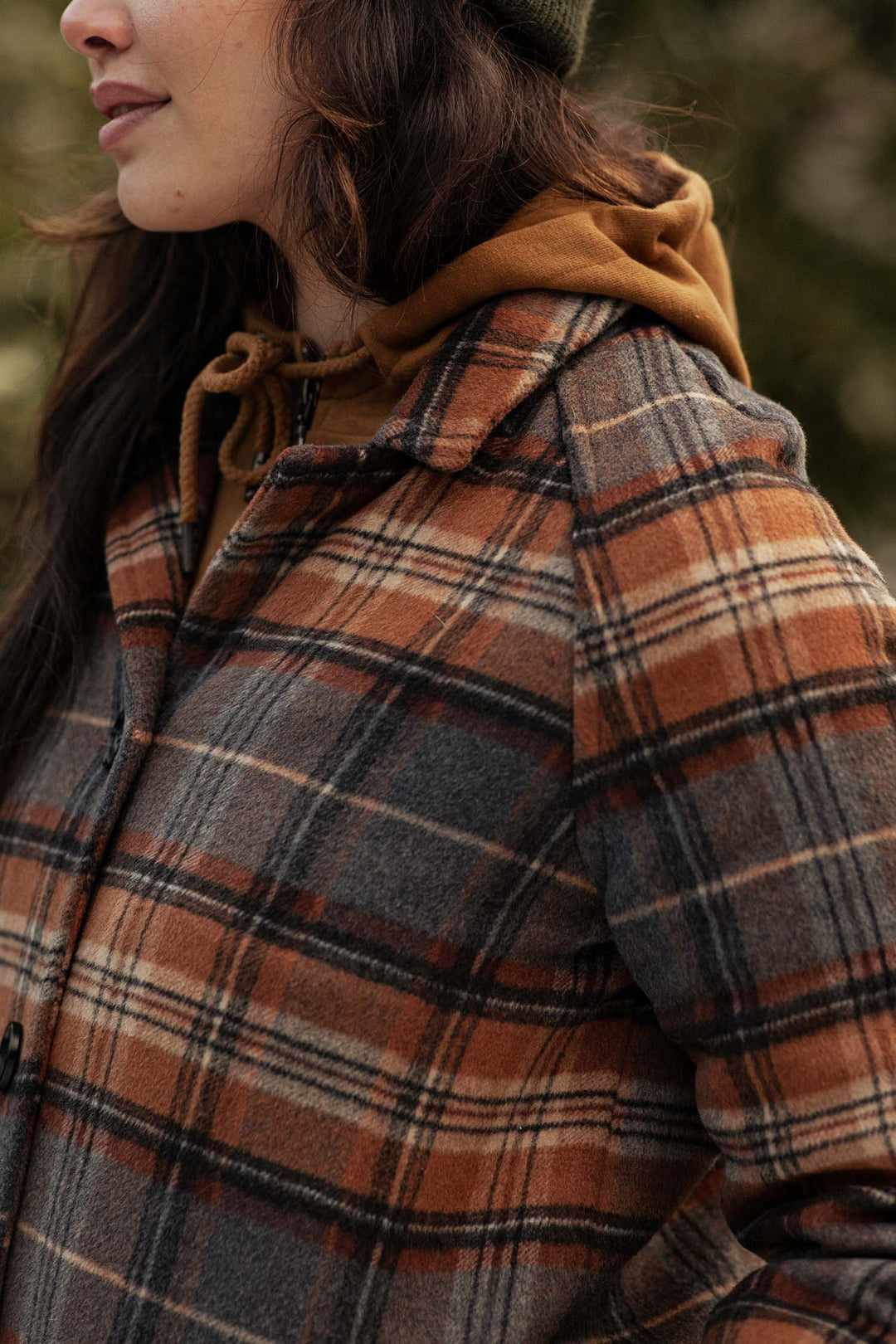 A person wears a brown hoodie under the Bridge & Burn Alberta Car Coat in Dorset Brushed Plaid—an orange, gray, and cream heavyweight wool blend. Standing outdoors, only the lower half of their face and upper body are visible.