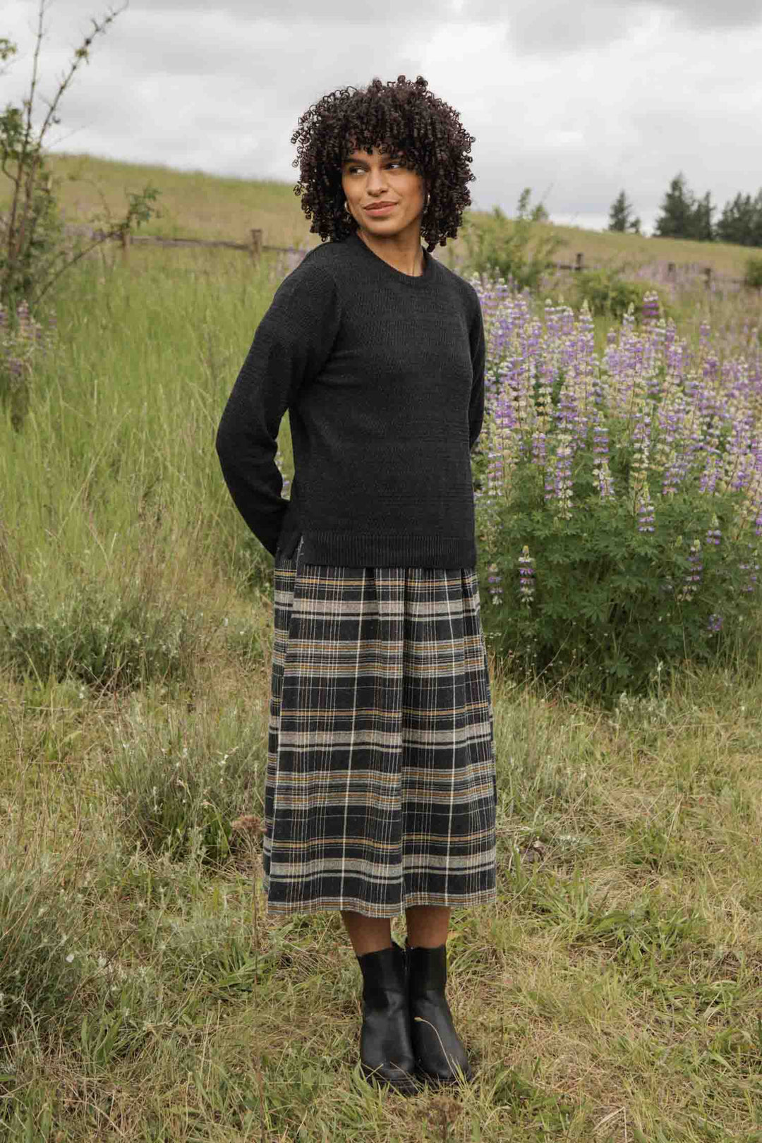 A woman with curly hair stands in a grassy field wearing the Bridge & Burn Ansel Sweater in Midnight. She pairs it with a plaid midi skirt and black ankle boots amid purple wildflowers and a wooden fence under cloudy skies.