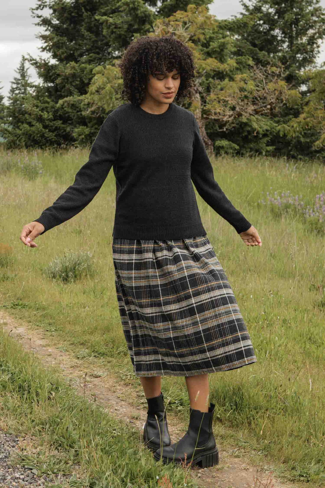 A woman with curly hair stands outdoors on a grassy path, wearing the Bridge & Burn Ansel Sweater in Midnight, paired with a plaid skirt and black boots. Trees and greenery form the background.
