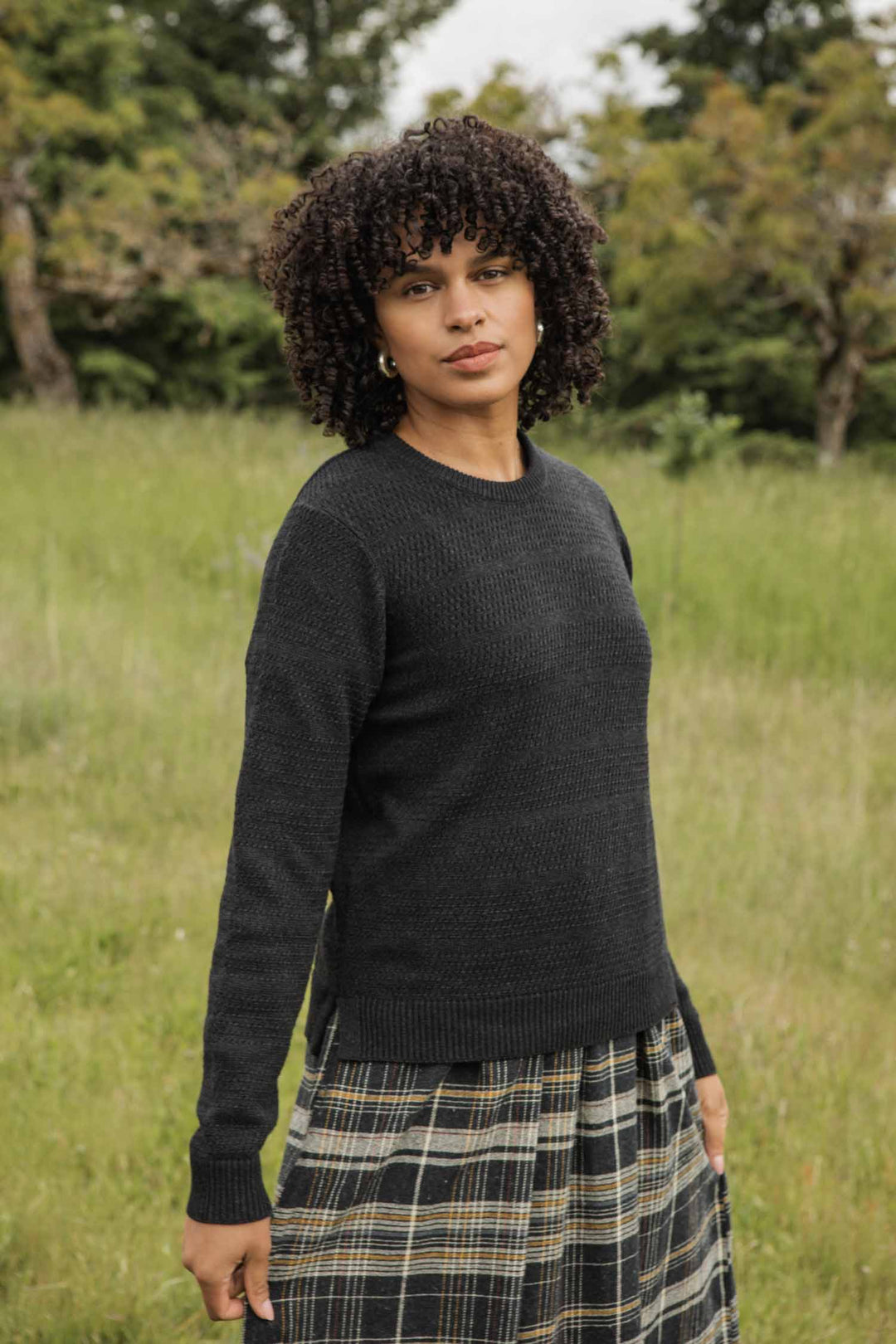 A woman with curly hair stands in a grassy field, wearing the Bridge & Burn Ansel Sweater in Midnight. Trees and greenery are visible behind her as she gazes at the camera with a neutral expression.