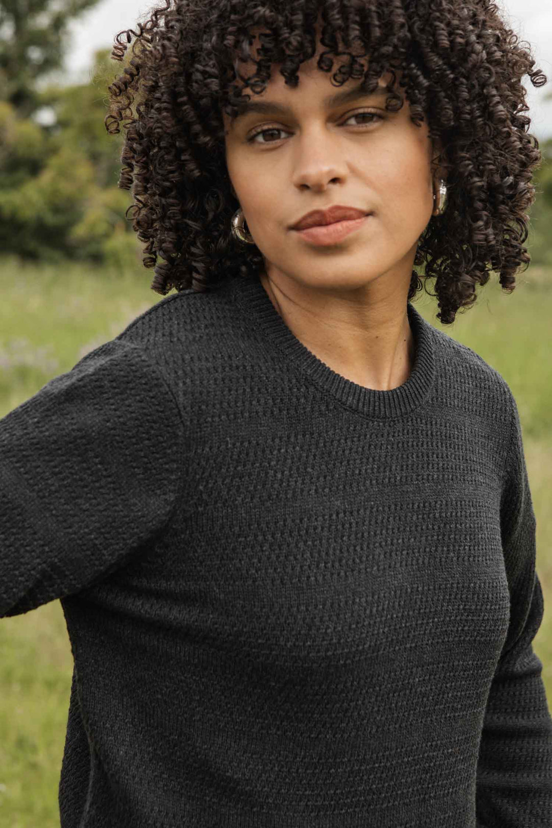 A woman with curly dark hair wears the Ansel Sweater in Midnight by Bridge & Burn. She stands outdoors in a grassy area, calmly looking at the camera with trees and blurred greenery in the background.