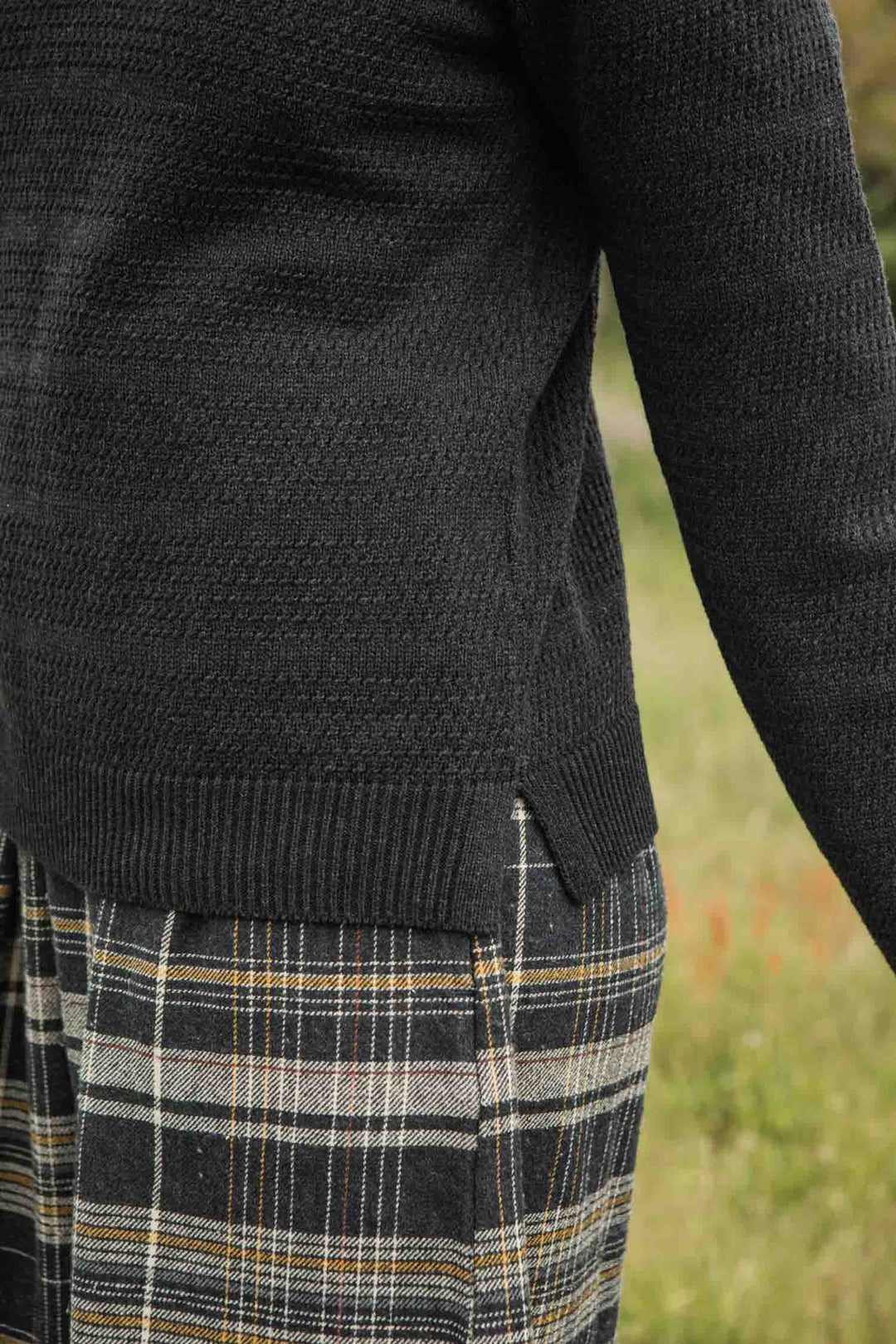 A person stands outside on grass, their side and arm partially visible, wearing the Bridge & Burn Ansel Sweater in Midnight paired with plaid pants.