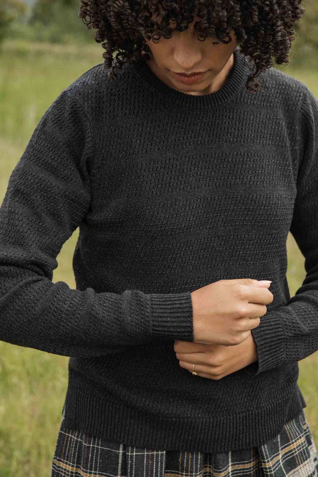 Wearing the Bridge & Burn Ansel Sweater in Midnight, a person with curly hair adjusts their sleeve outdoors in a grassy area, paired with a plaid skirt.