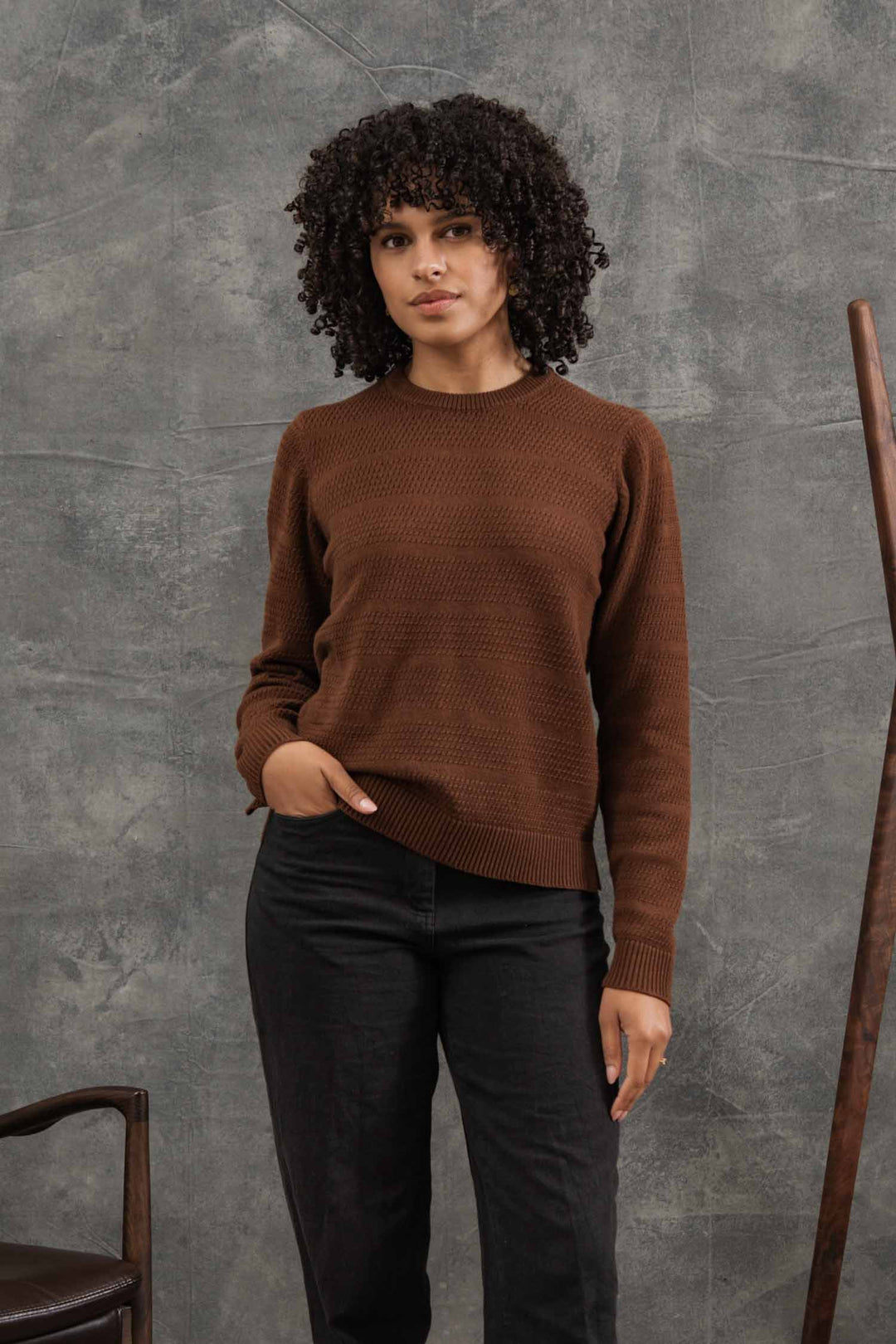 A person with curly hair models the Bridge & Burn Ansel Sweater in Walnut, a mid-weight brown sweater made from recycled fibers, paired with black pants and posed indoors against a textured gray background—a sustainable essential.