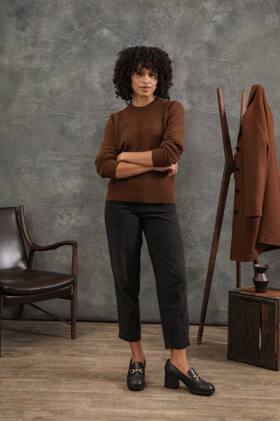 A woman with curly hair wears the Bridge & Burn Ansel Sweater in Walnut, paired with black pants and heeled loafers. She stands with arms crossed in a room featuring a brown chair, coat stand, and brown coat against a textured gray backdrop.
