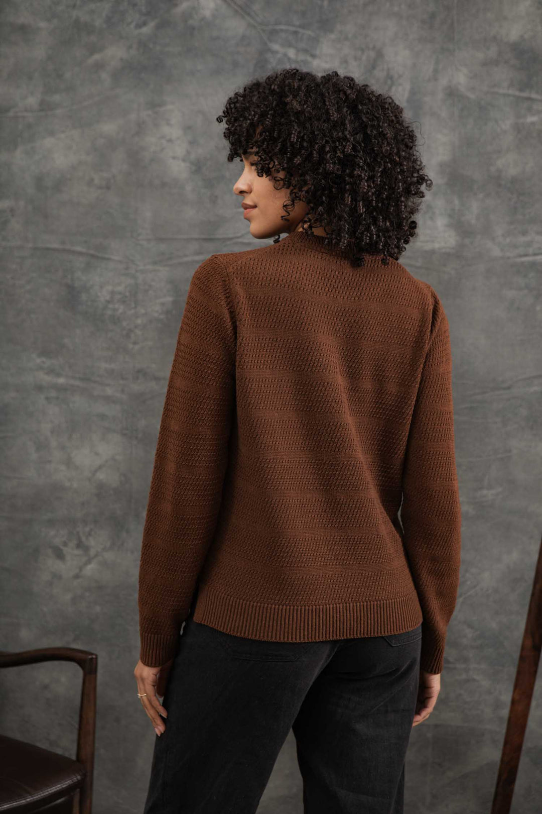 A person with curly hair is seen from behind, wearing the Bridge & Burn Ansel Sweater in Walnut and black pants, set against a textured gray backdrop.
