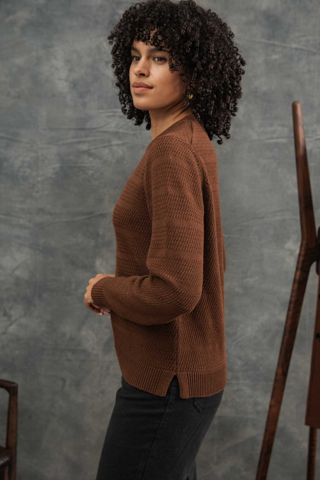 A woman with curly hair stands in profile against a gray textured background, wearing the Bridge & Burn Ansel Sweater in Walnut and black pants. A wooden object is partially visible on the right.