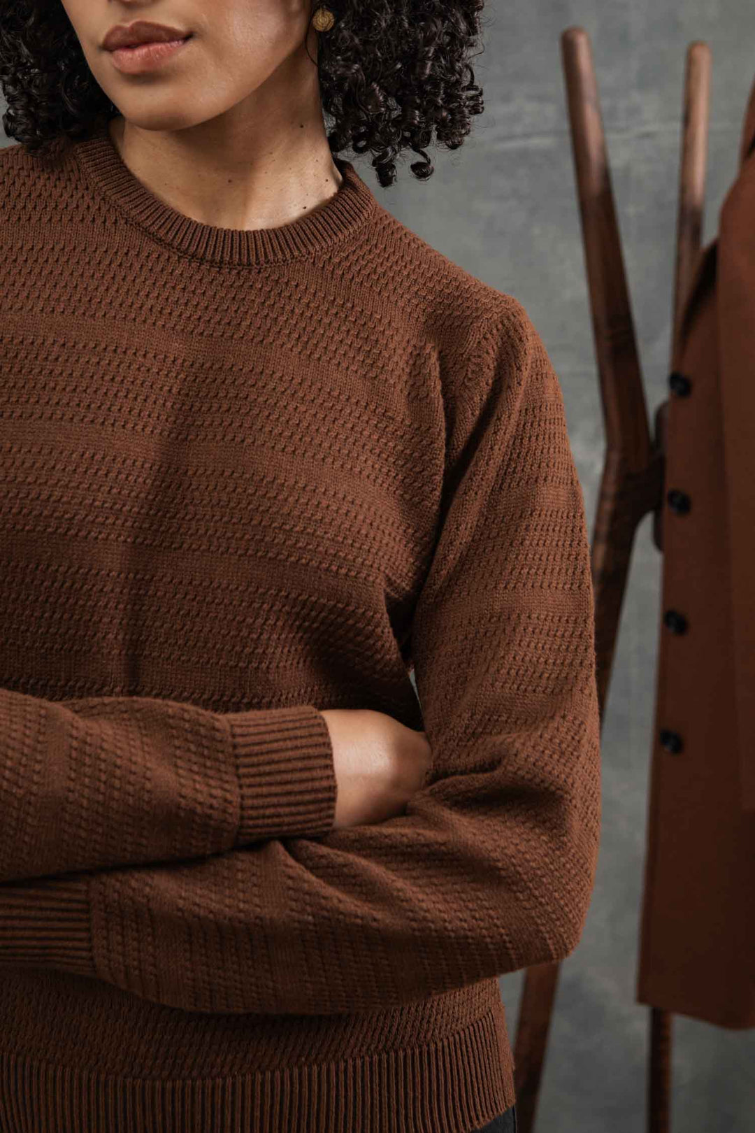 A person with curly hair stands with arms crossed, wearing the Ansel Sweater in Walnut by Bridge & Burn—a sustainable, mid-weight brown sweater crafted from recycled fibers. A wooden coat rack and brown coat are blurred in the background.