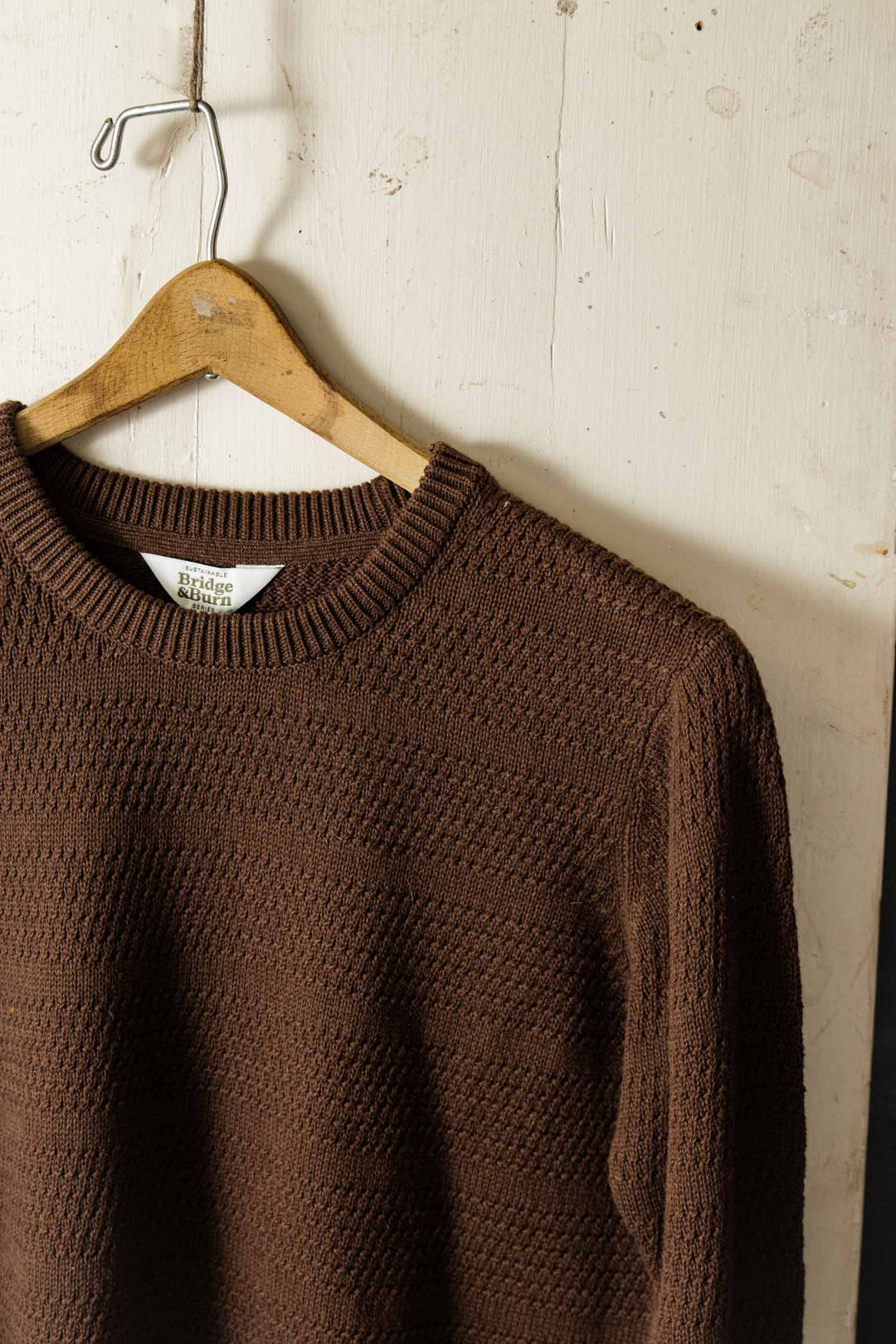 The Ansel Sweater in Walnut by Bridge & Burn—a brown, textured mid-weight sweater made from recycled fibers—hangs on a wooden hanger against a slightly worn white wall, showcasing its sustainable design.
