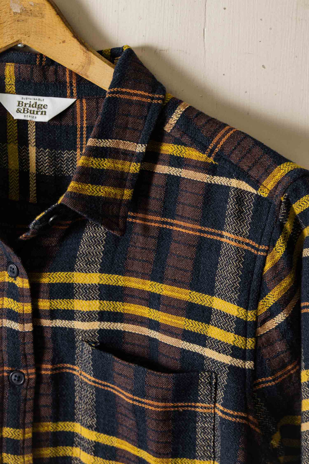 A close-up of the Bridge & Burn Ash Slim Shirt in Desert Dawn Plaid hanging on a wooden hanger. This organic cotton shirt features dark blue, yellow, brown, and cream stripes with a collar, partial button placket, and chest pocket.
