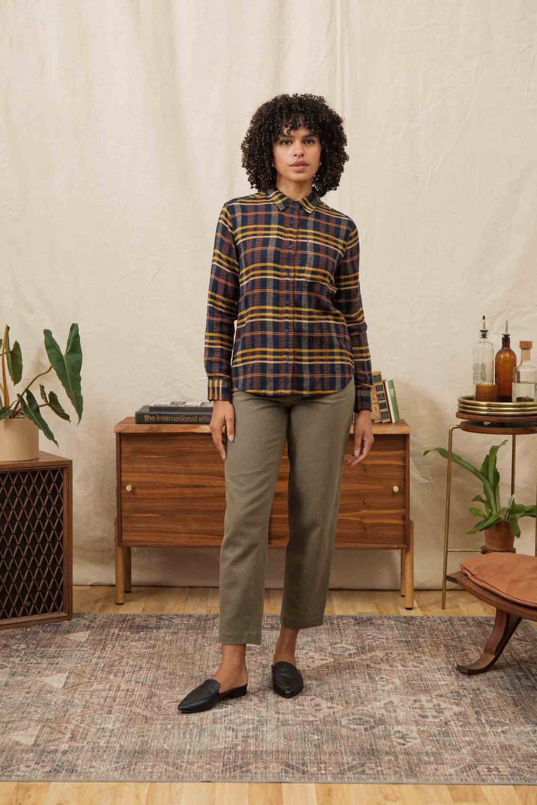 A woman stands indoors on a patterned rug, wearing the Bridge & Burn Ash Slim Shirt in Desert Dawn Plaid, olive green pants, and black loafers. A wooden console with books, plants, and bottles is behind her.