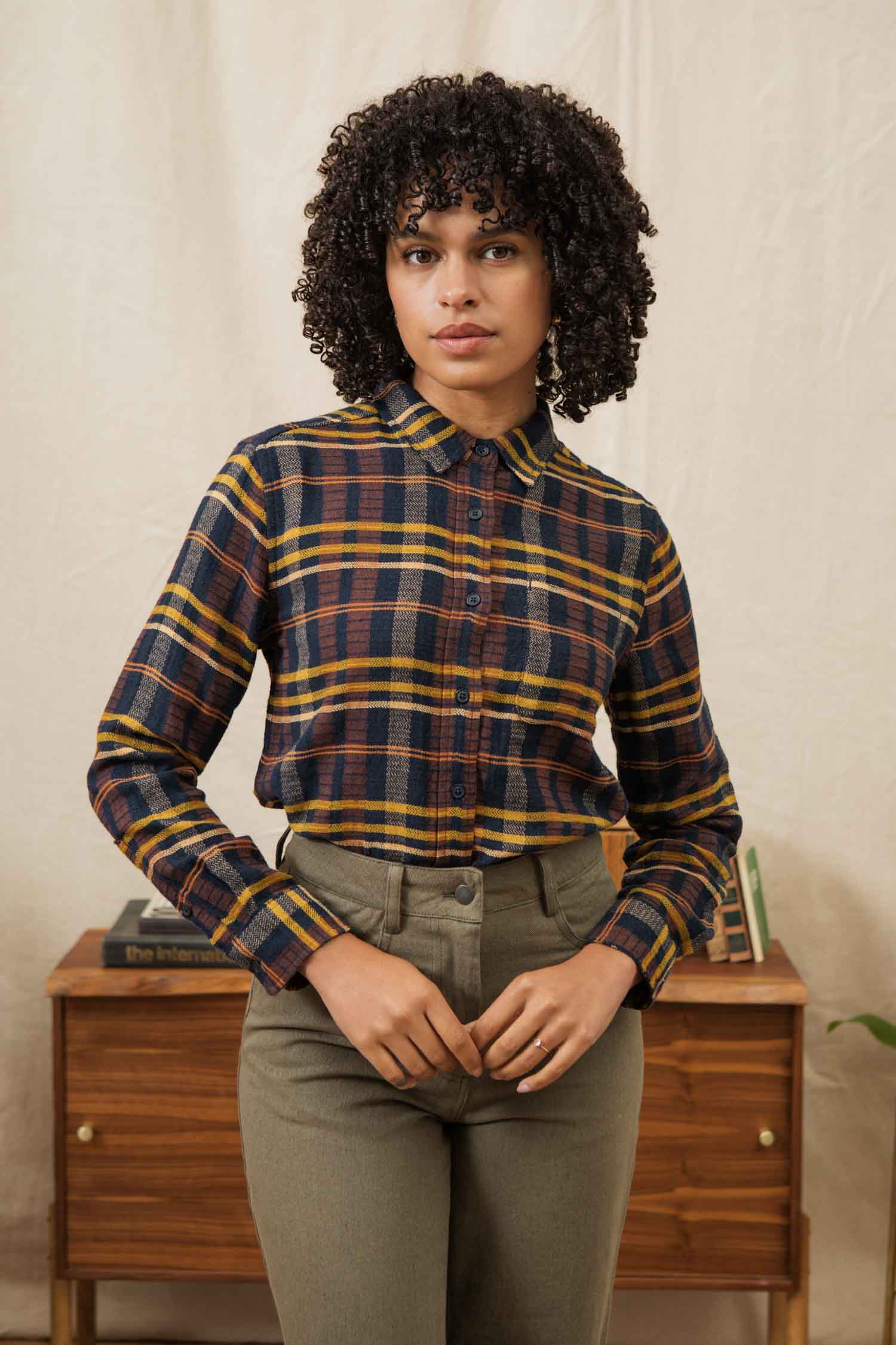 A person with curly hair wears the Bridge & Burn Ash Slim Shirt in Desert Dawn Plaid and high-waisted pants, standing before a wooden table with books and a plant, set against a light-colored background.